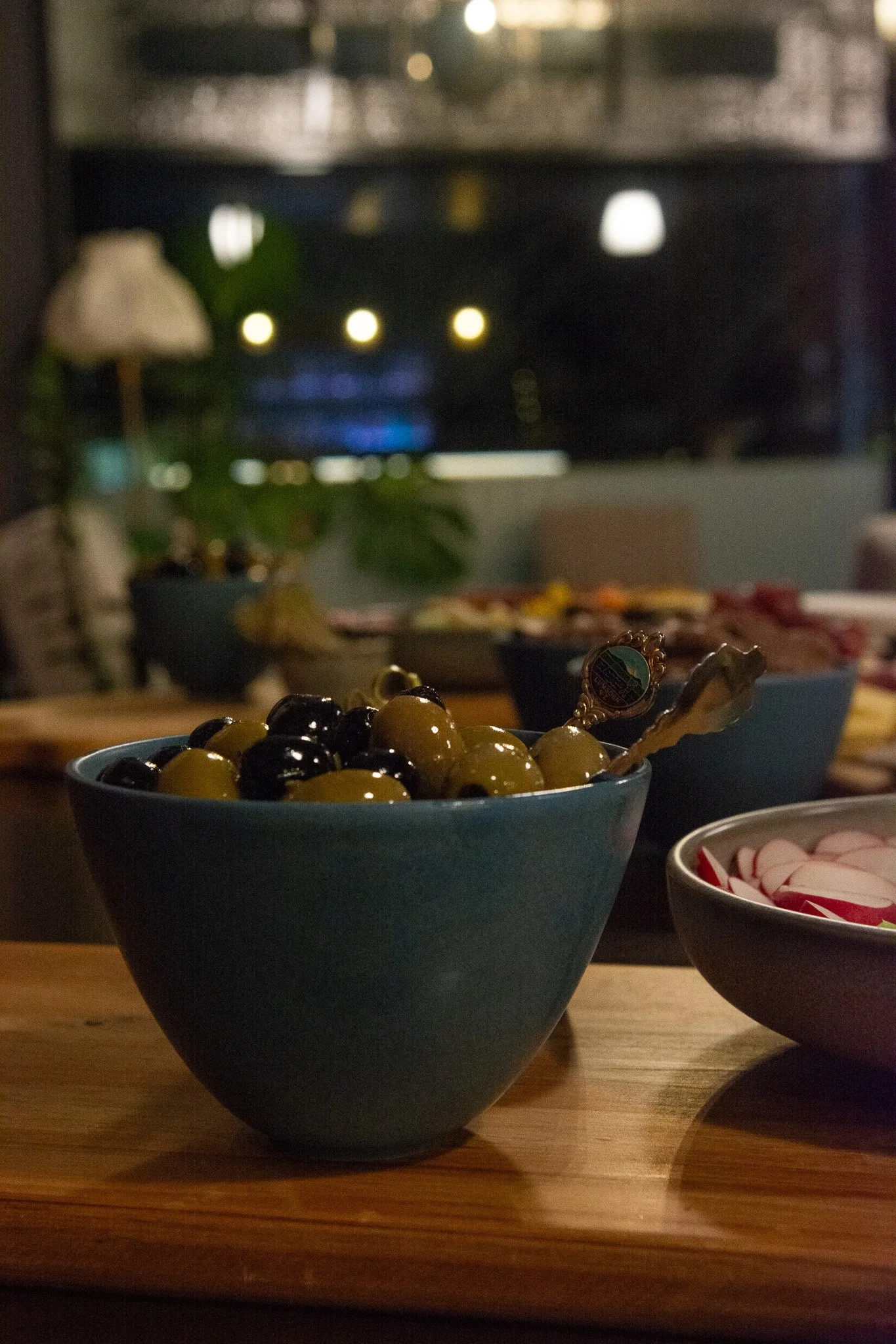 olives in blue ceramic bowl with radish crudite, on wooden board with window and lamp in background