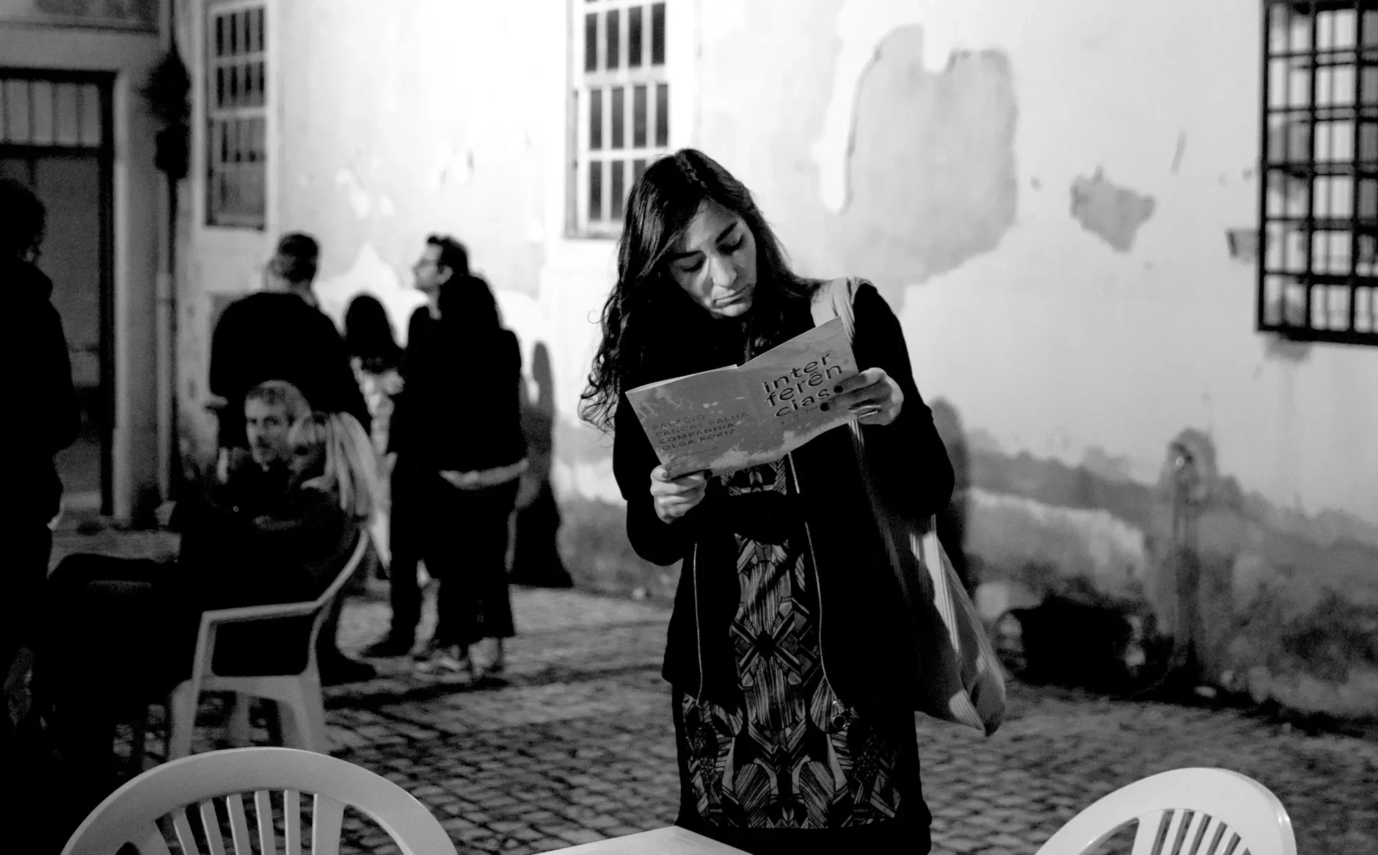 Pessoa jovem lendo um jornal em uma rua de noite, com um grupo de pessoas ao fundo