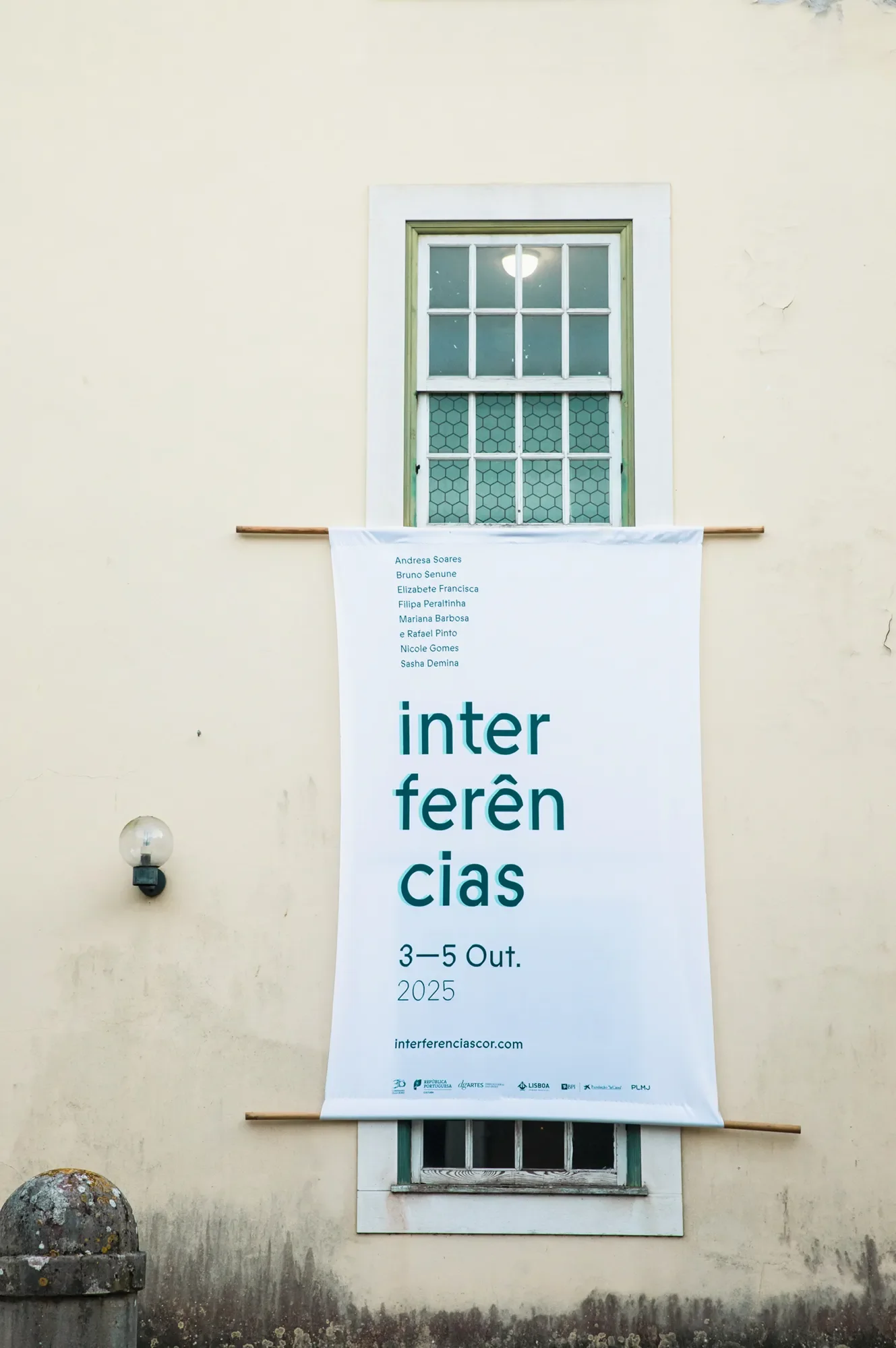 Banner publicitário de um evento chamado 'Interferências' com datas de 3 a 5 de outubro de 2025, e uma lista de nomes de pessoas na parte superior, fixado na parede de uma casa com janela de vidro. A parede da casa é de cor clara e há uma luminária ao lado esquerdo do banner.