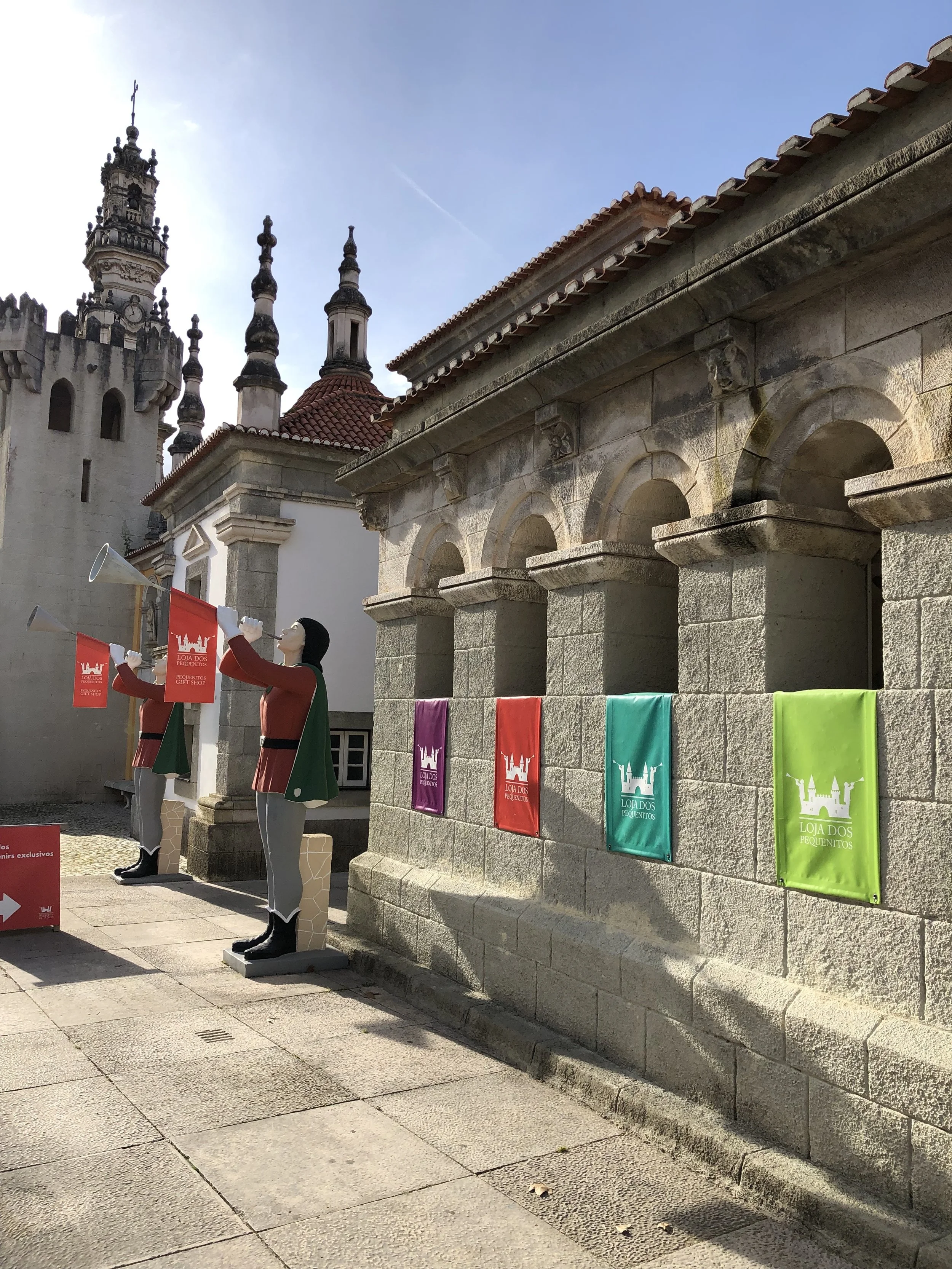 Estátuas de figuras humanas vestidas com roupas coloridas, segurando trompetes, colocadas ao lado de um muro de pedra com bandeiras coloridas e um castelo antigo ao fundo.
