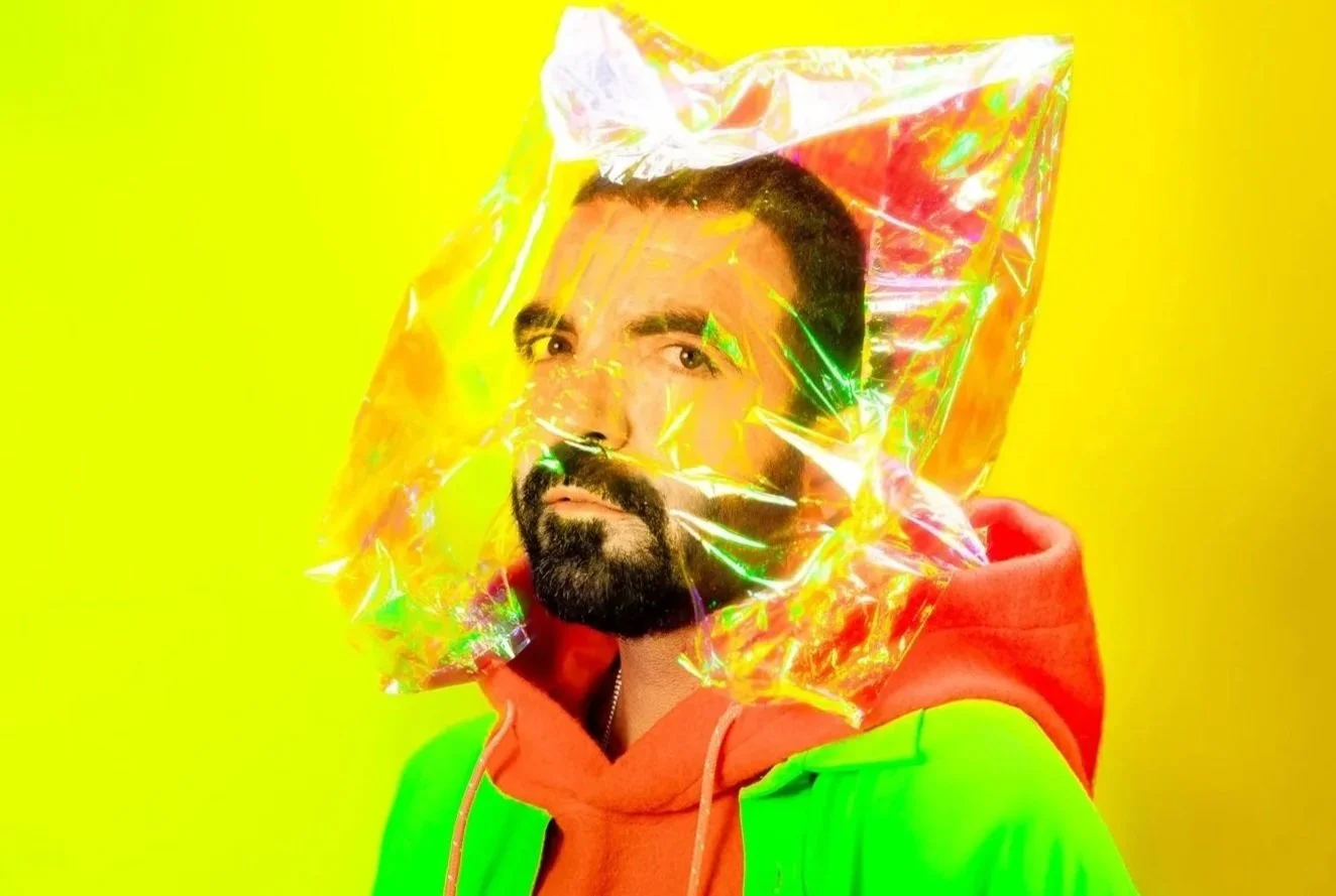 Homem com barba e cabelo curto usando jaqueta verde e capuz laranja, com uma capa de plástico holográfico na cabeça, contra fundo amarelo.