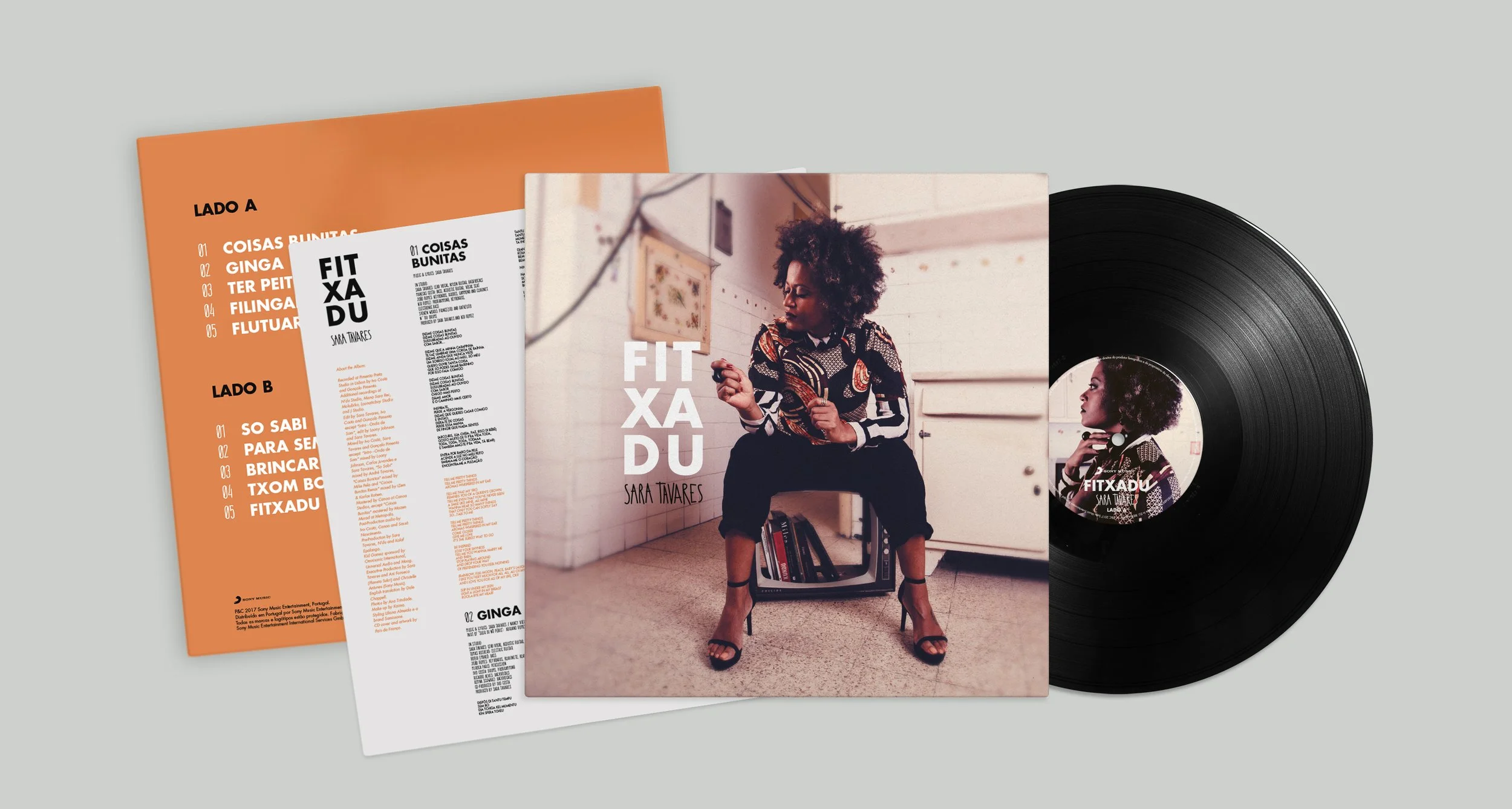 Capa de álbum com uma mulher sentada em uma cadeira, vestindo uma blusa de estampa colorida e calça preta, ao lado de um toca-discos e um livro, com uma parede branca ao fundo. Ao lado, uma vitrola preta e uma folha com informações do álbum, incluindo uma lista de faixas.