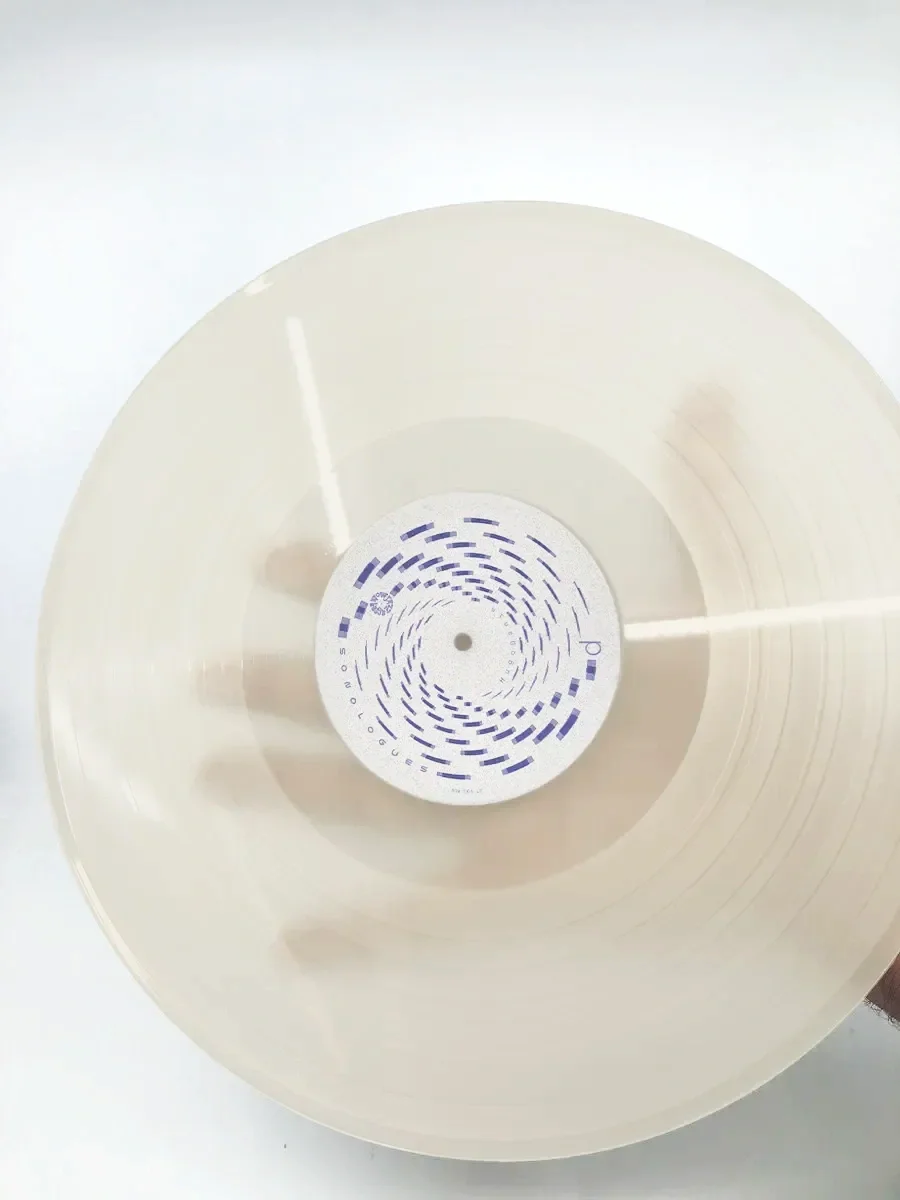 Vista superior de um vinil de 12 polegadas com etiqueta ao centro, mostrador de um disco de vinil de cor clara com design gráfico e texto em azul.