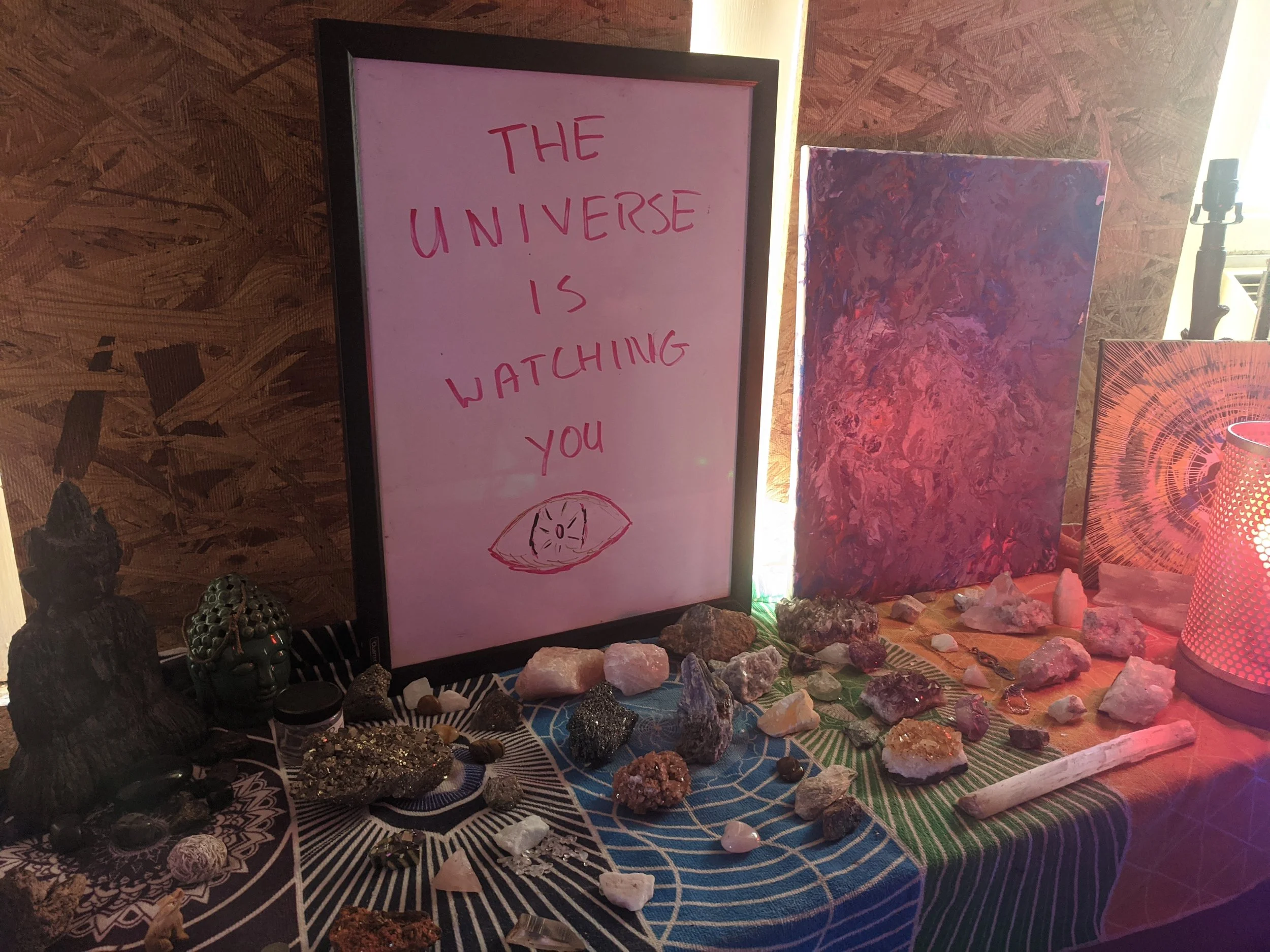 A table with a pink framed sign that reads 'The universe is watching you' with a drawing of an eye, various crystals, rocks, and small statues, including a Buddha head, arranged on it. There is a purple and orange abstract painting and a patterned lampshade in the background.