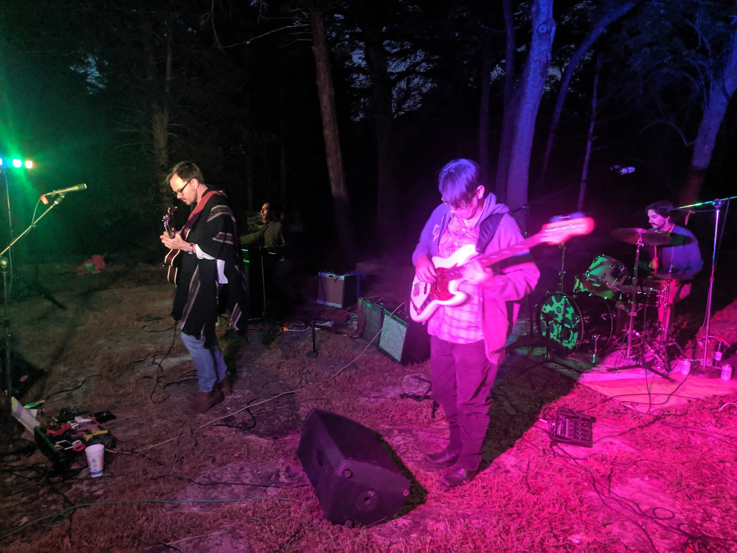 A band performs outdoor at night with purple and green lighting, including a guitarist, bassist, and drummer, surrounded by trees.