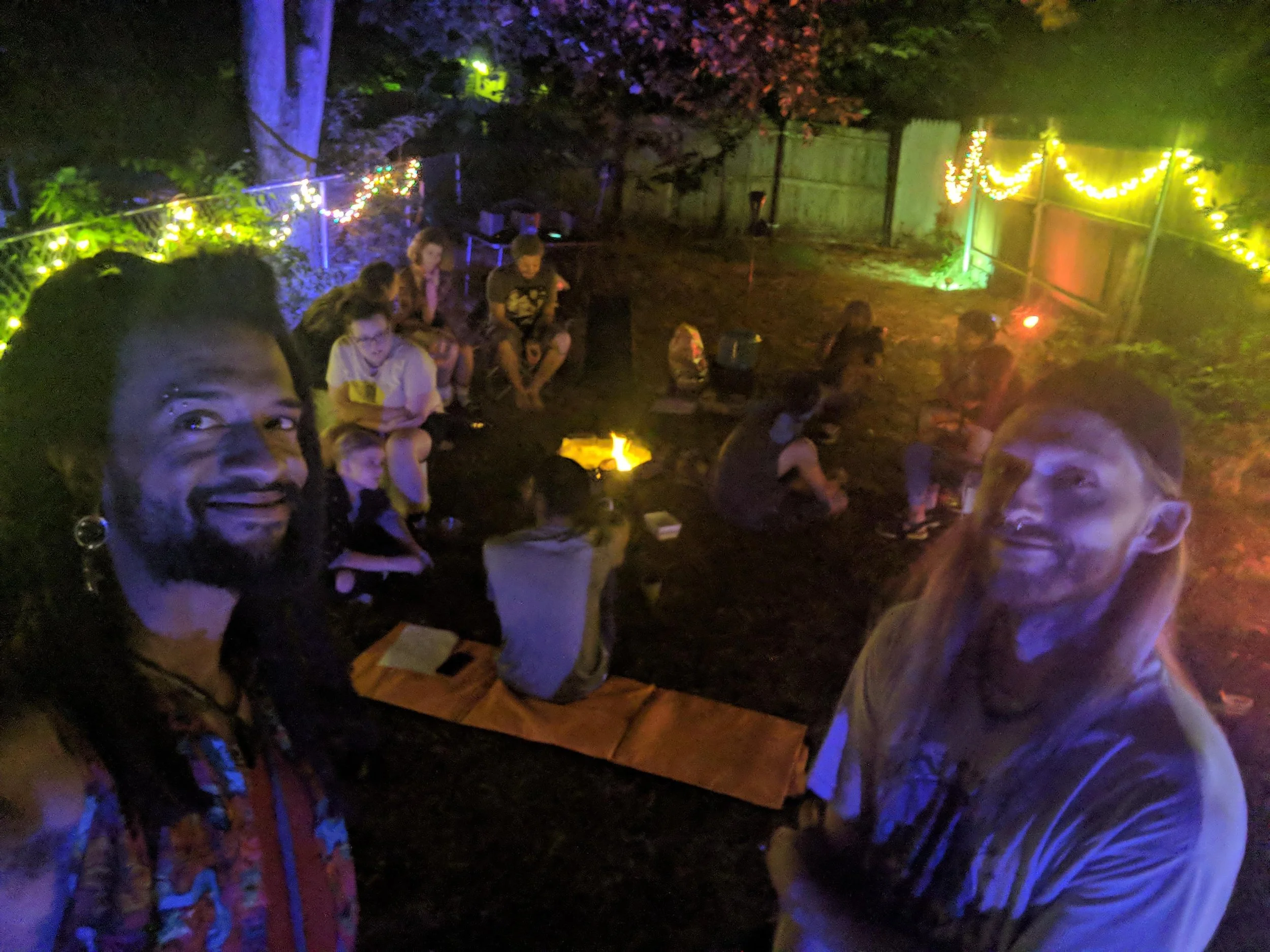 People gathered outdoors at night around a small fire, with colorful string lights illuminating the area, possibly during a backyard gathering or party.