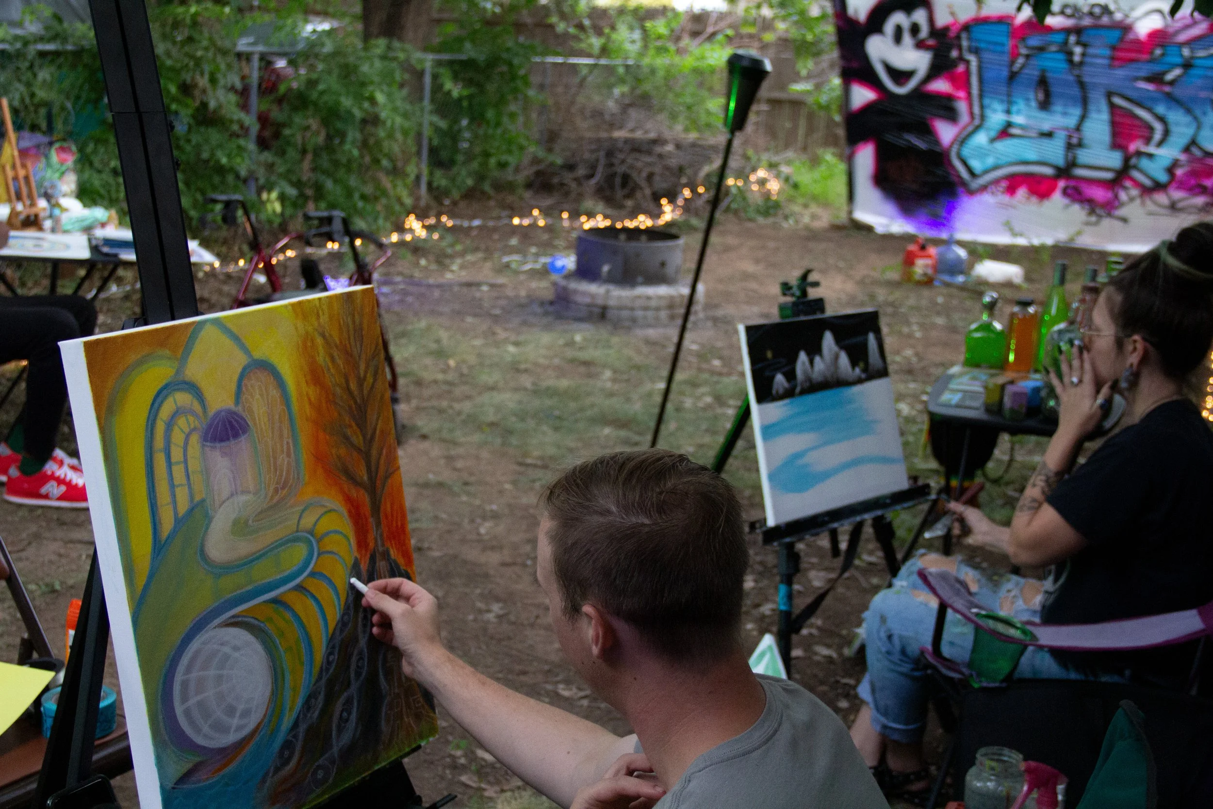 People painting colorful artwork outdoors in a backyard with fairy lights and graffiti on a backdrop.
