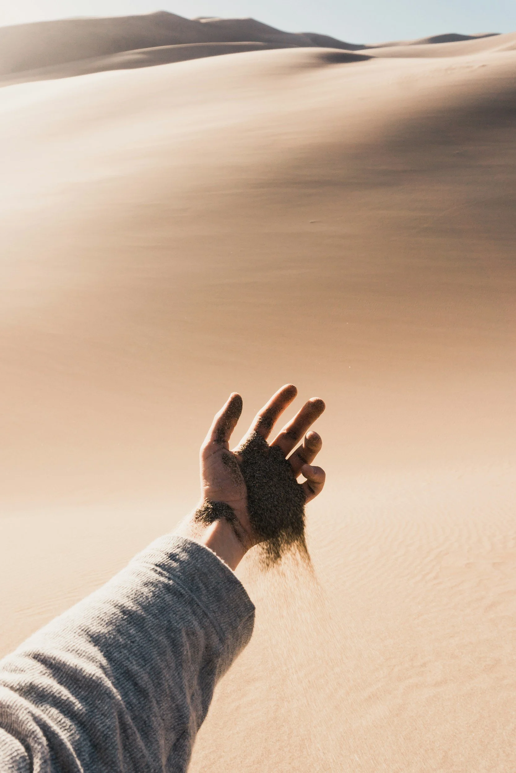A hand releasing sand into a desert landscape with sand dunes in the background.