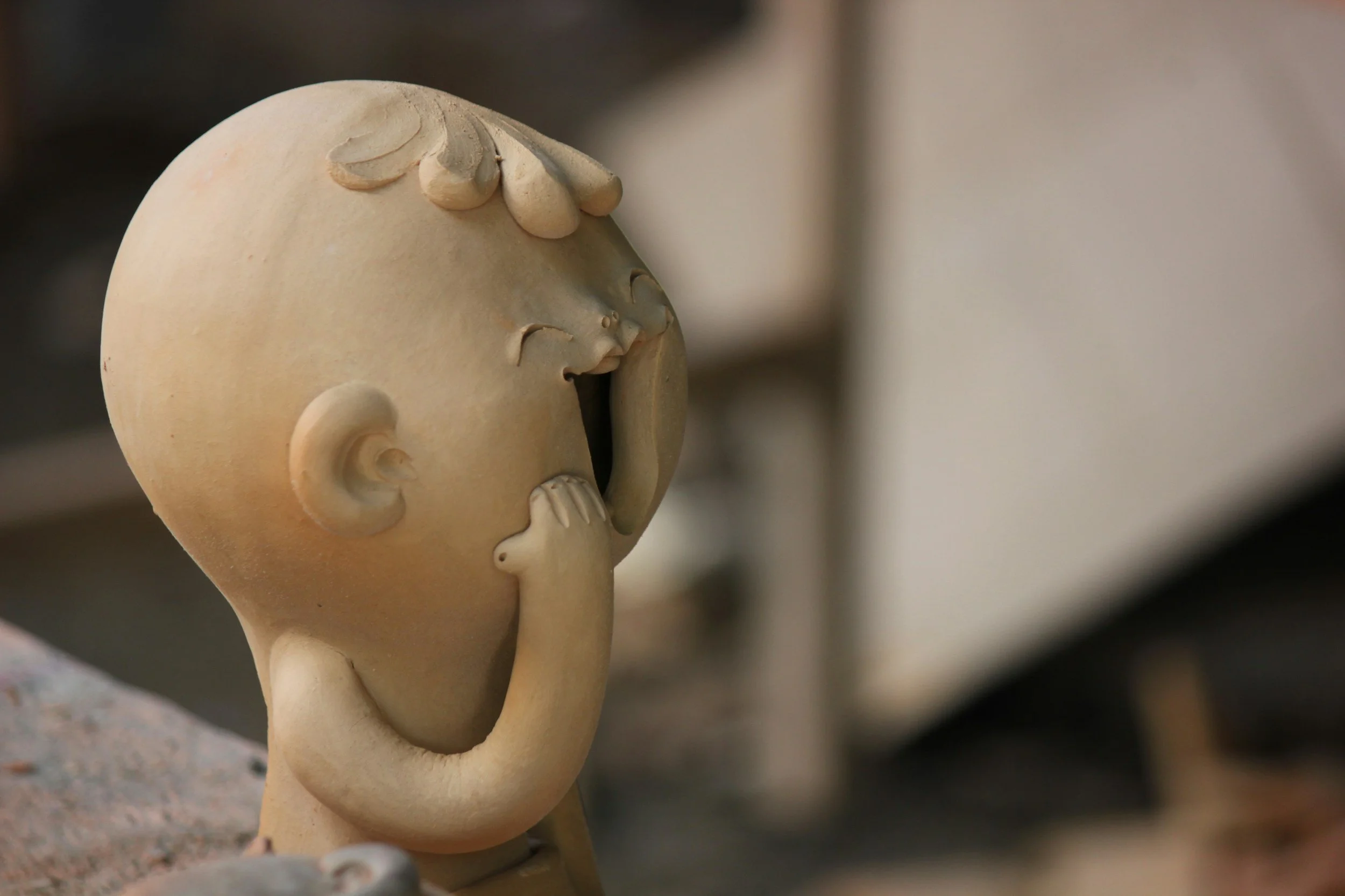 A ceramic sculpture of a child with an open mouth, closed eyes, and hand on cheek, with a dreamy expression, in a neutral indoor setting.