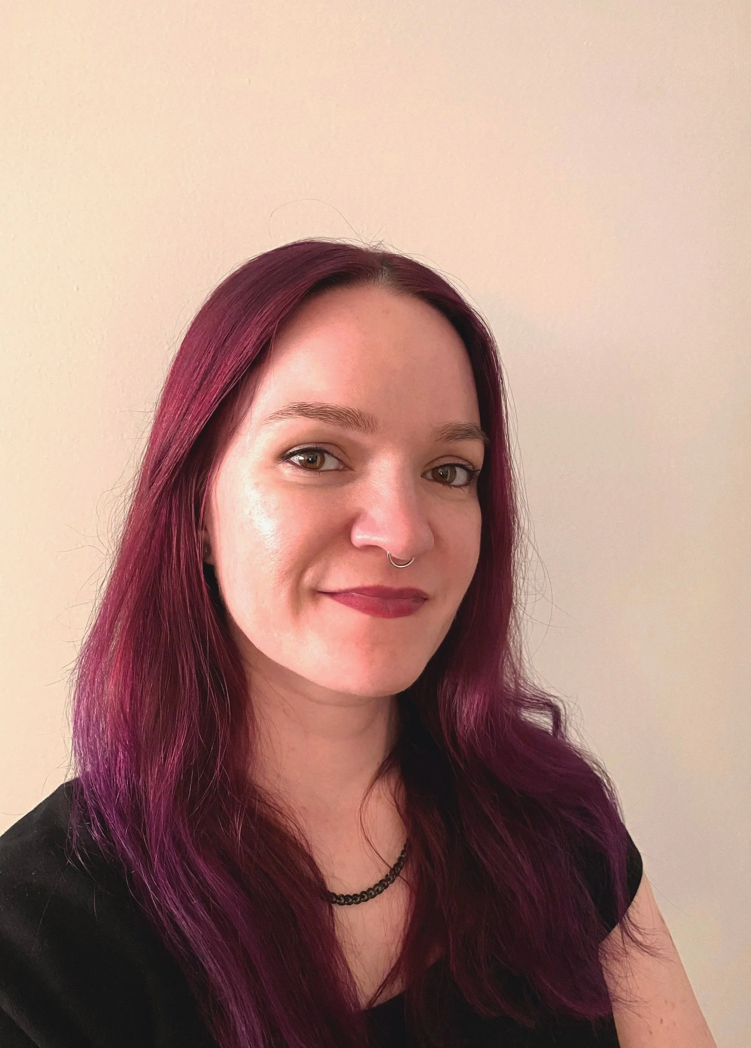 A woman with purple hair, wearing a black shirt, a black necklace, and a septum piercing, smiling in front of a plain beige wall.