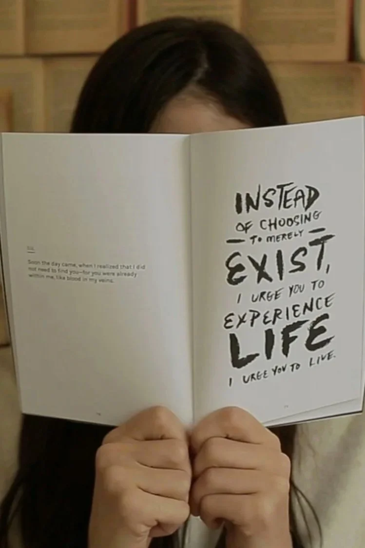 A person with dark hair holding an open book to conceal their face. The right page of the book has a handwritten quote in large, bold letters that reads, 'Instead of choosing to merely exist, I urge you to experience life. I urge you to live.' The background appears to be a wooden wall or paneling.