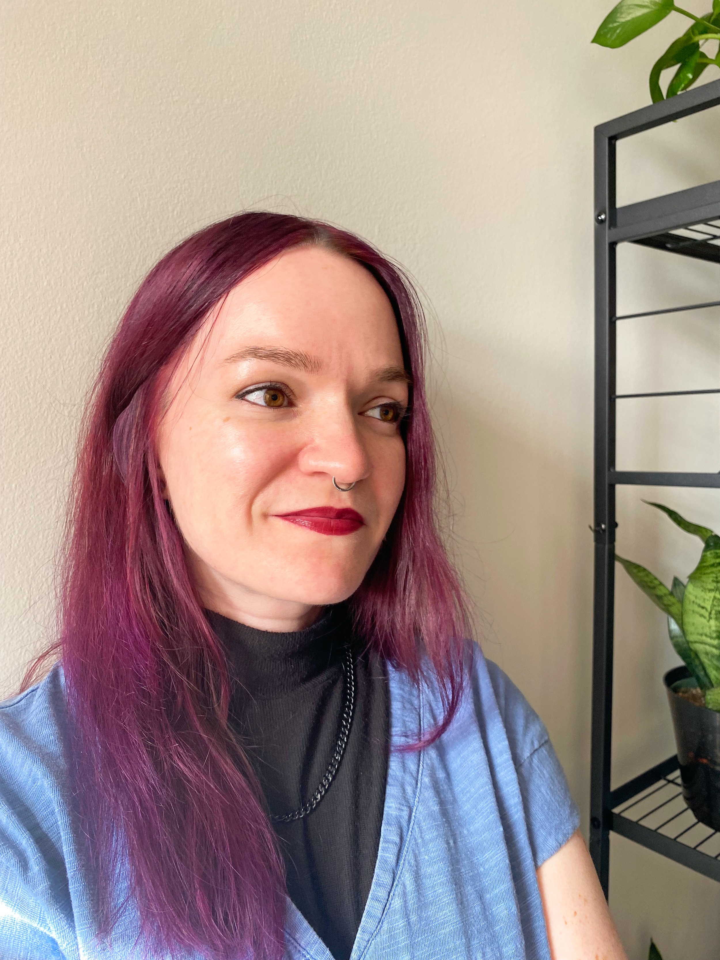 A woman with shoulder-length purple hair and a septum piercing, wearing a black top and blue scrubs, sitting near a black metal shelf with potted plants against beige walls.