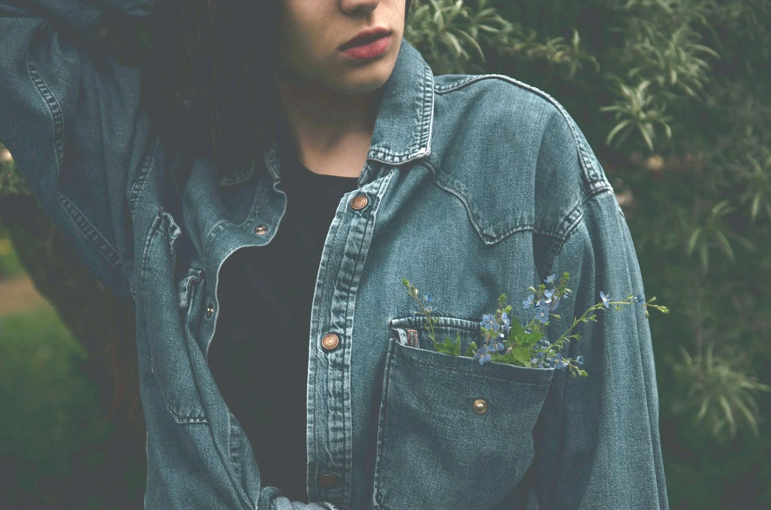 Close-up of a person wearing a denim jacket with a pocket containing blue flowers, standing outdoors with green foliage in the background.