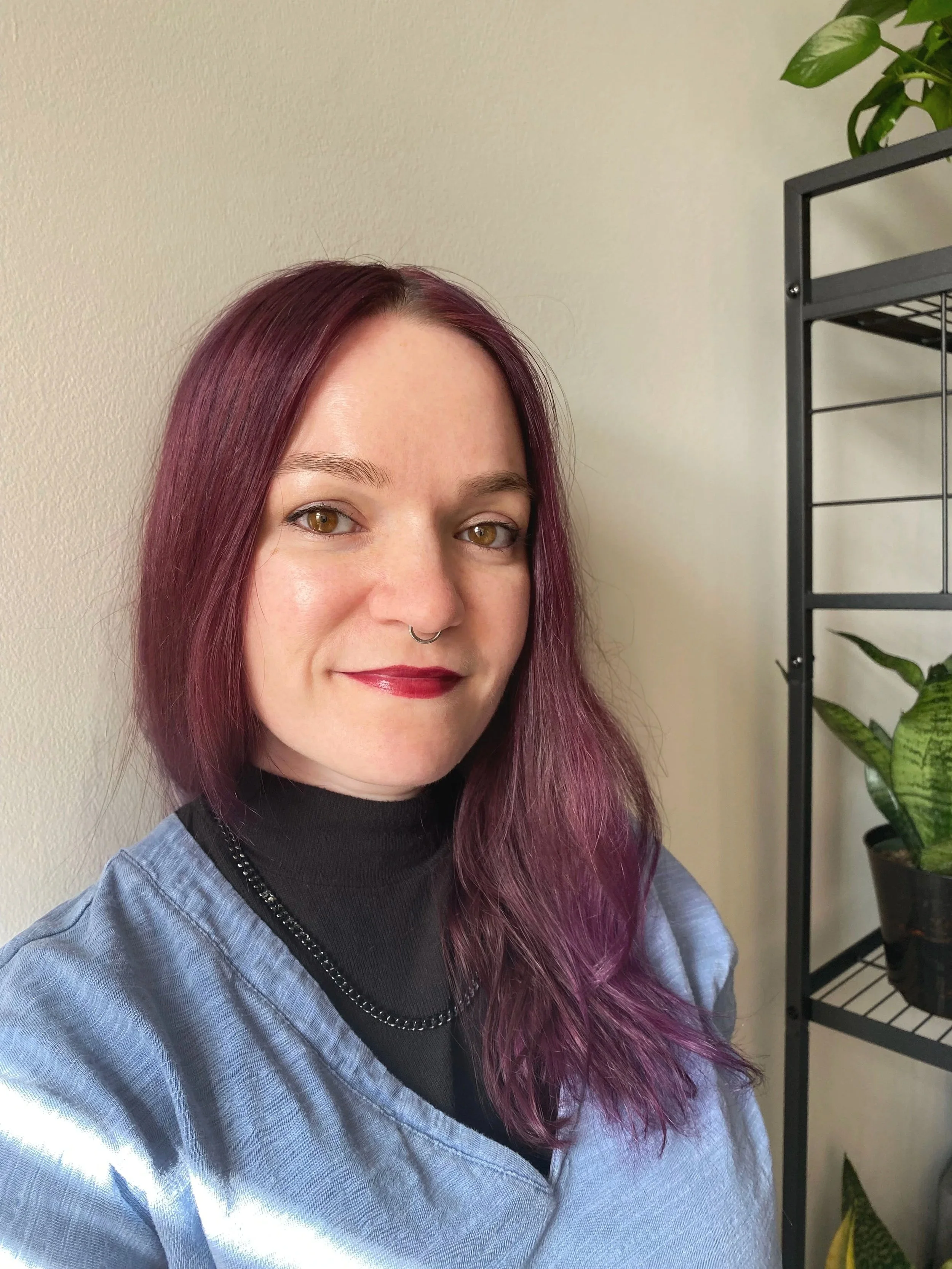 A woman with medium-length purple hair, wearing a black turtleneck and a blue scrub top, taking a selfie indoors with a plant and a black metal shelf in the background.