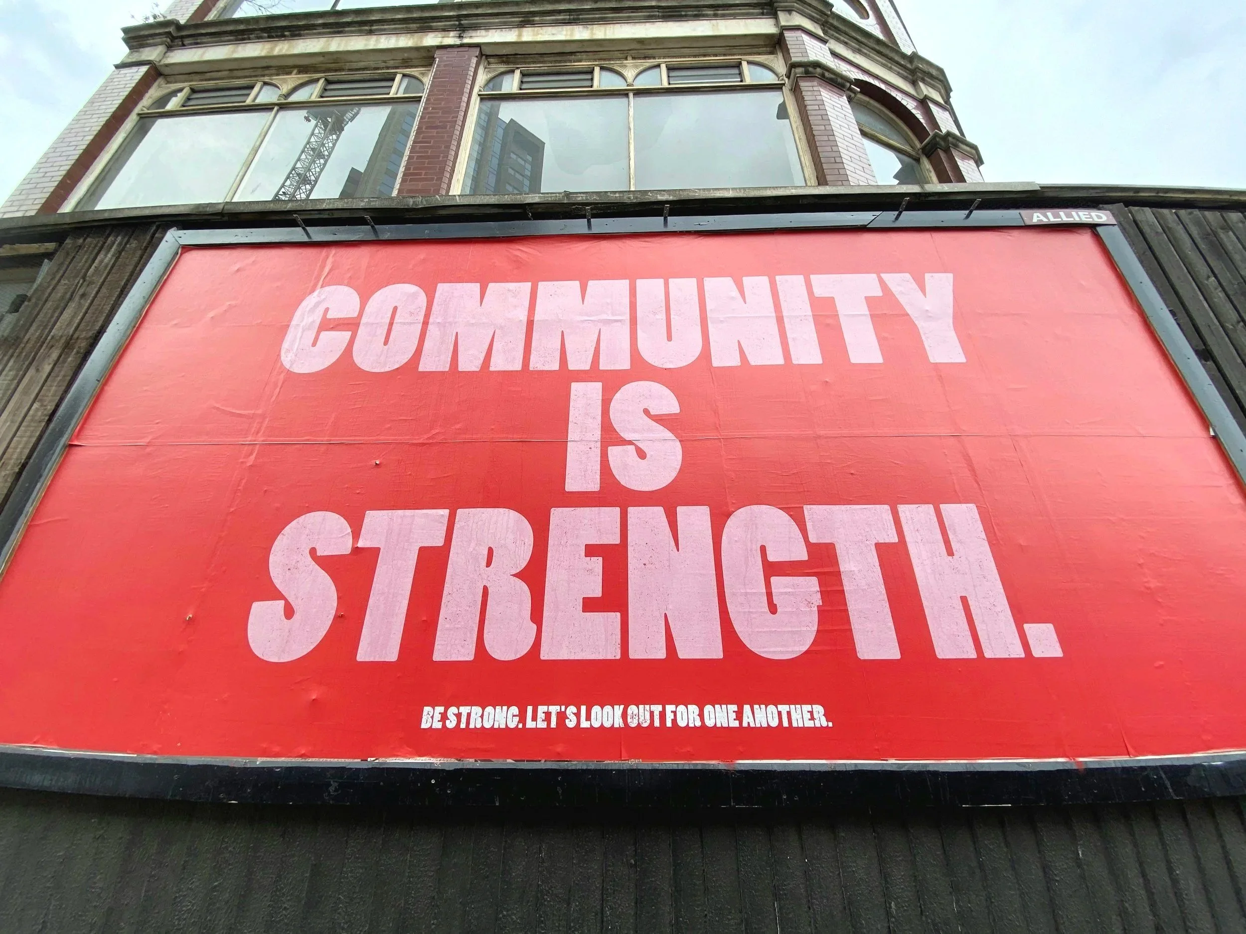 A large red billboard with the message "Community is Strength" and smaller text that reads "Be strong. Let's look out for one another." The billboard is located on a building with tall windows and a tower crane is visible in the background.