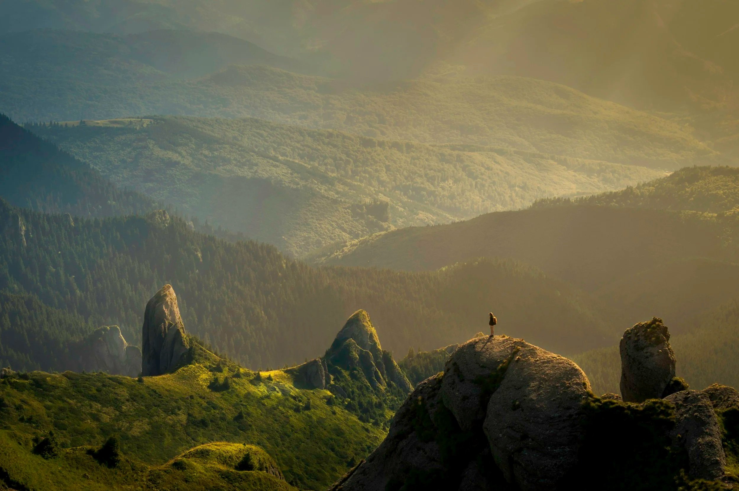 A person standing on a large rock formation overlooking a series of mountains covered in dense forests during sunset or sunrise with sunlight casting a golden hue over the landscape.