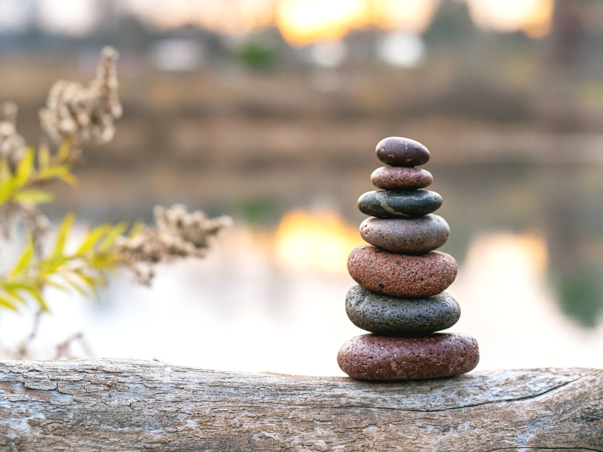A stack of smooth, rounded stones balanced on a piece of driftwood near the water with a blurred background of a water body and sunset sky.