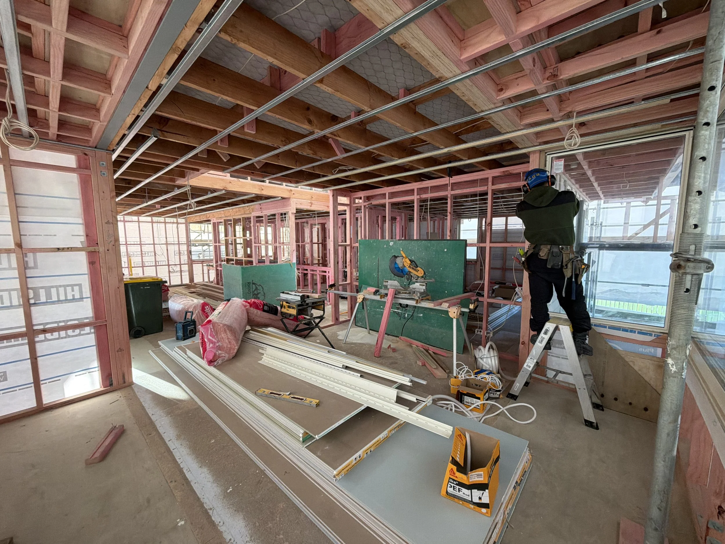 Construction worker installing windows frames in a partially built house, with exposed wooden and metal framing, tools, materials, and insulation on the floor.