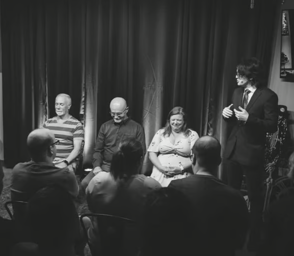 A black and white photo of a magic performance with a group of four people sitting on stage and one magician standing and performing to the audience, in front of a small seated audience.