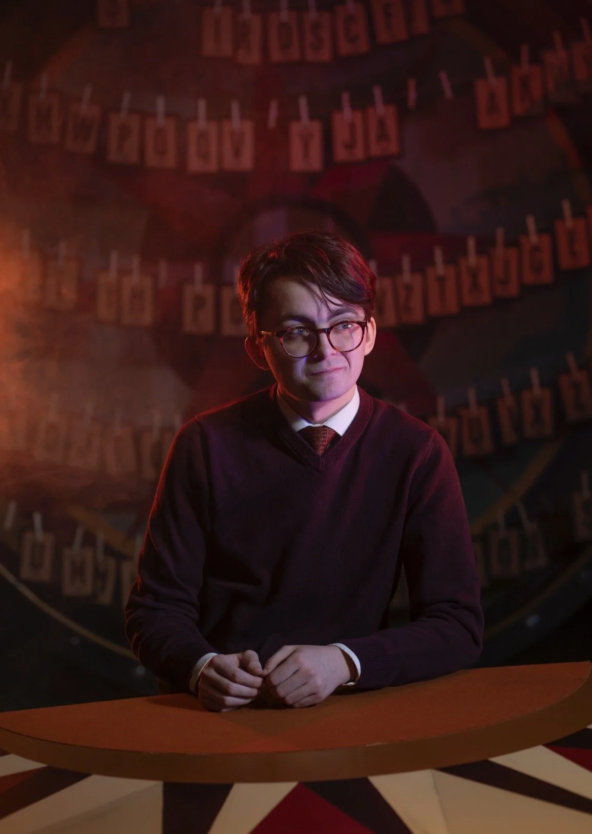 A young man with dark hair, glasses, and a serious expression, dressed in a dark sweater and a shirt with a tie, sitting at a wooden table in a dimly lit room. The background features a prop with letters and small photographs hanging around it.
