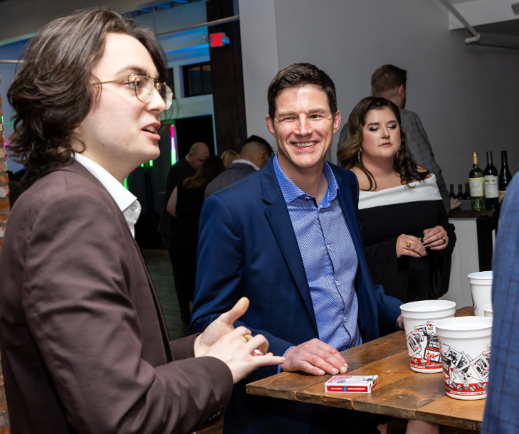Three people standing around a wooden table at a social gathering. The man in the center is smiling and wearing a blue blazer and light blue shirt. The woman on the right is dressed in black and white, holding a cup, and the man on the left has shoulder-length brown hair, glasses, and is wearing a brown blazer. There are menus or placemats and cups on the table, and bottles of wine and glasses in the background.