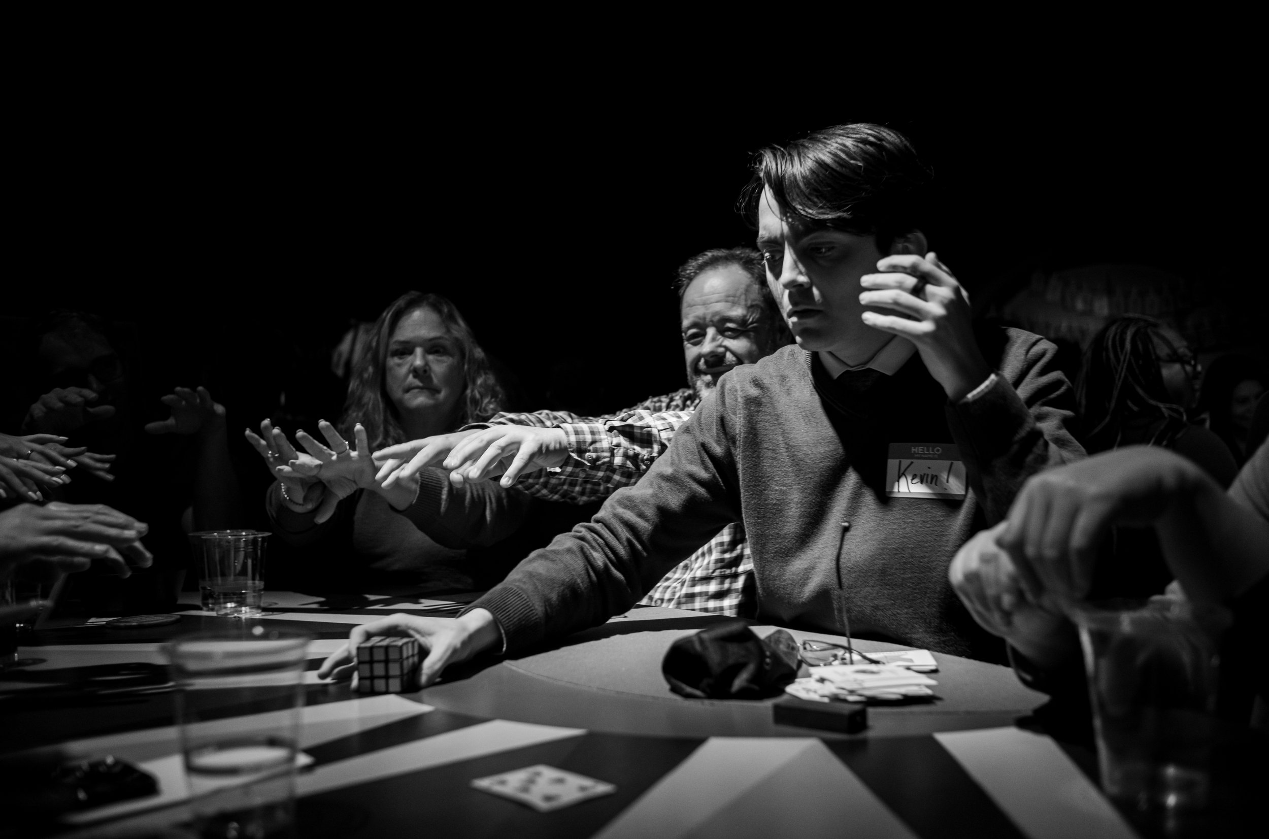People at a table watching a magician in foreground reaching out with a Rubik's Cube, surrounded by others reaching and observing.