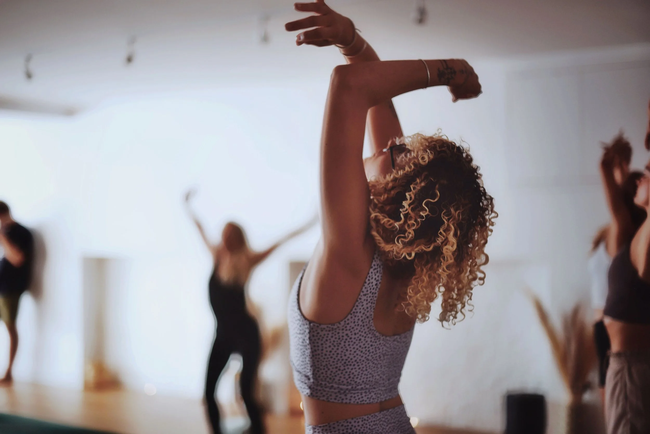 A woman with curly hair practicing yoga in a studio, surrounded by other women also engaging in yoga or stretching exercises.