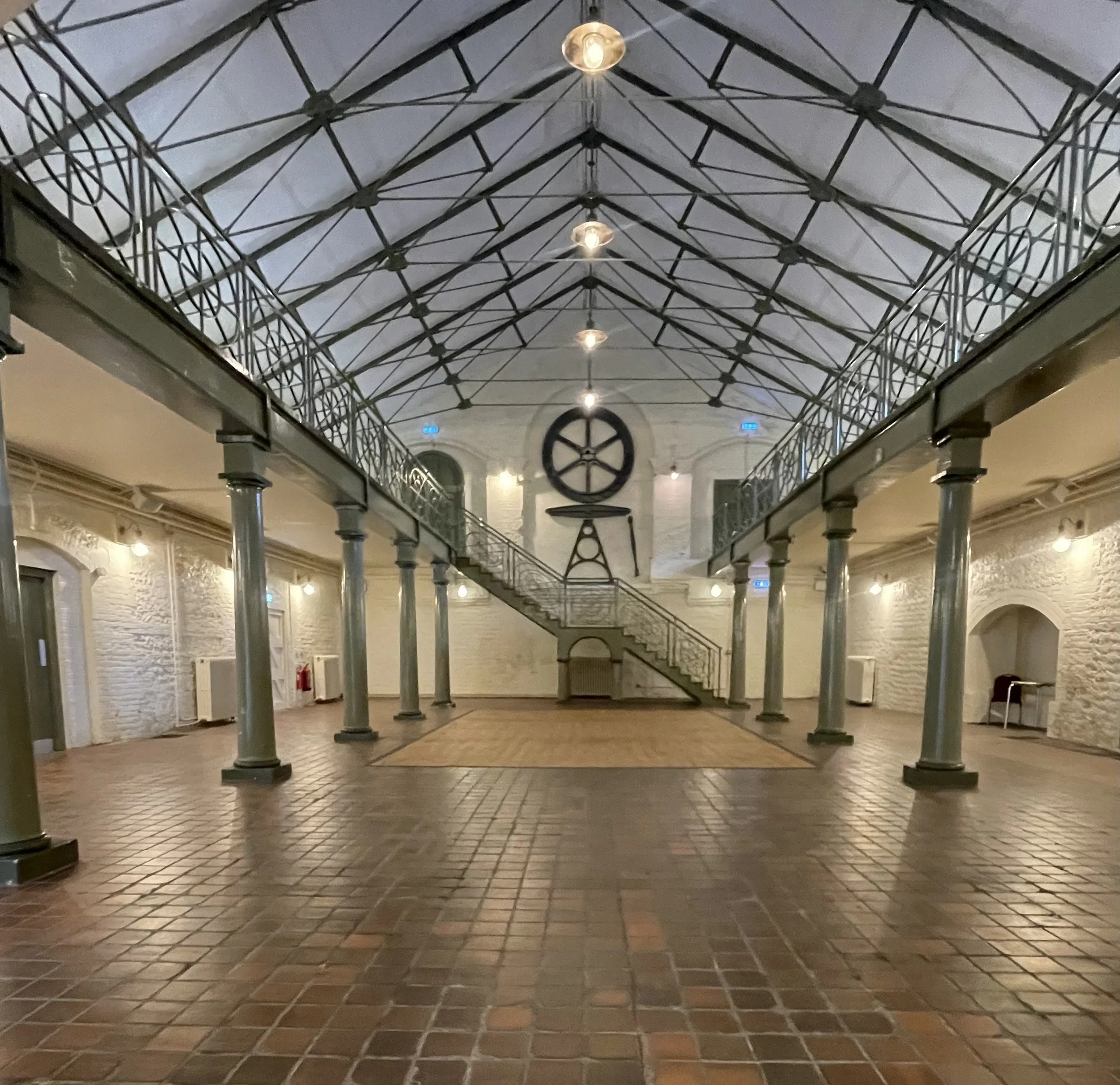 Interior of a spacious hall with brick flooring, white brick walls, metal columns, a staircase, and a high glass ceiling with hanging lights.