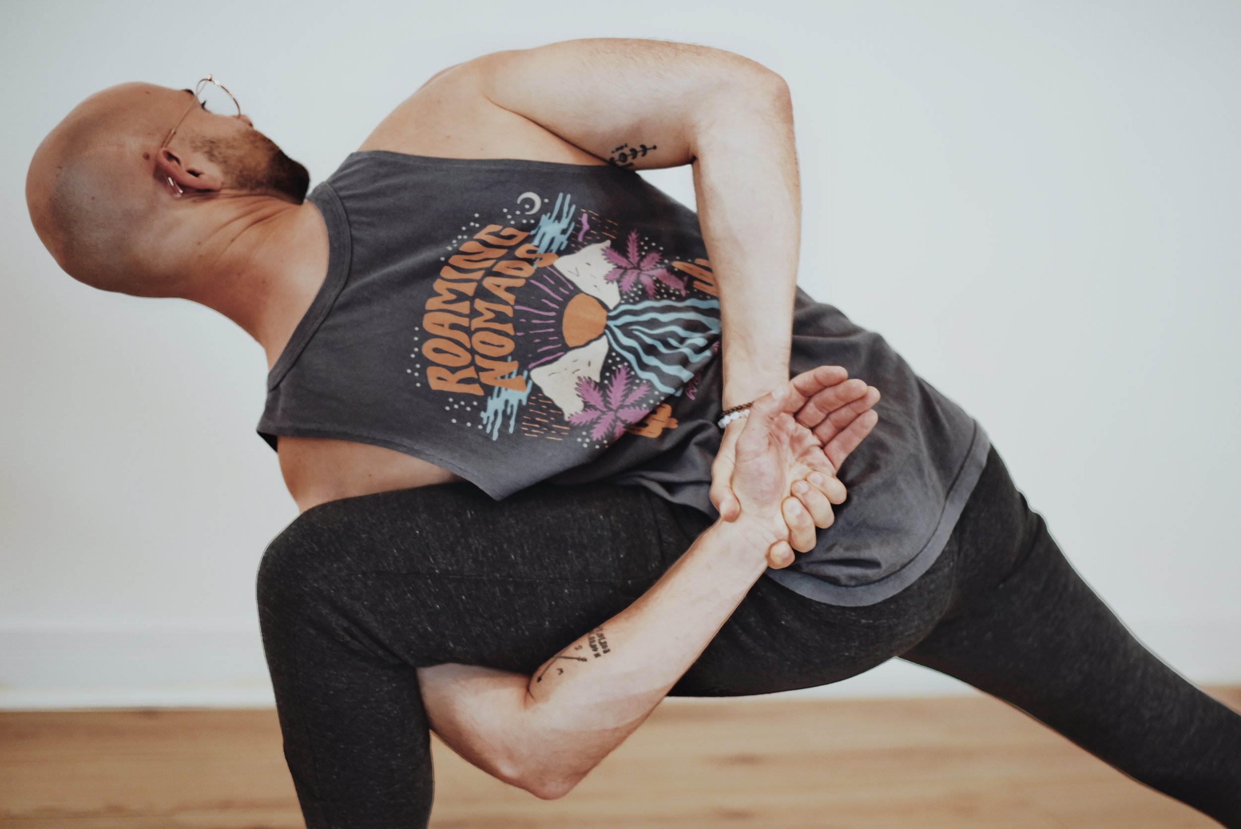 A person practicing yoga in a challenging pose, balancing on one leg while holding their foot with one hand and extending the other arm behind them. They are wearing a sleeveless shirt with a sunset and mountain design and dark leggings, indoors with a white wall background.