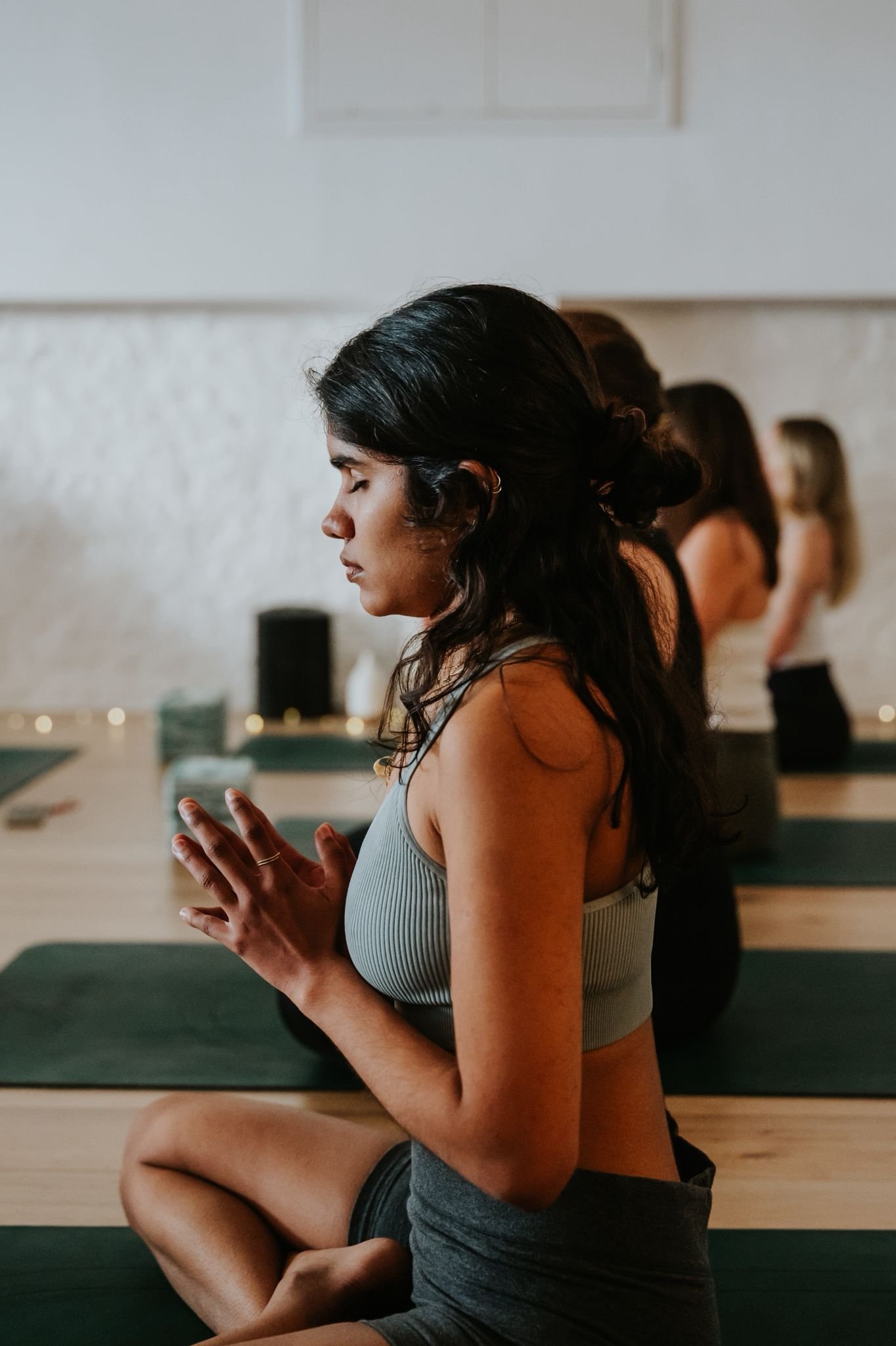 A woman practicing yoga in a meditation pose in a class, with her hands together and makeup serene expression, surrounded by other women in similar poses.