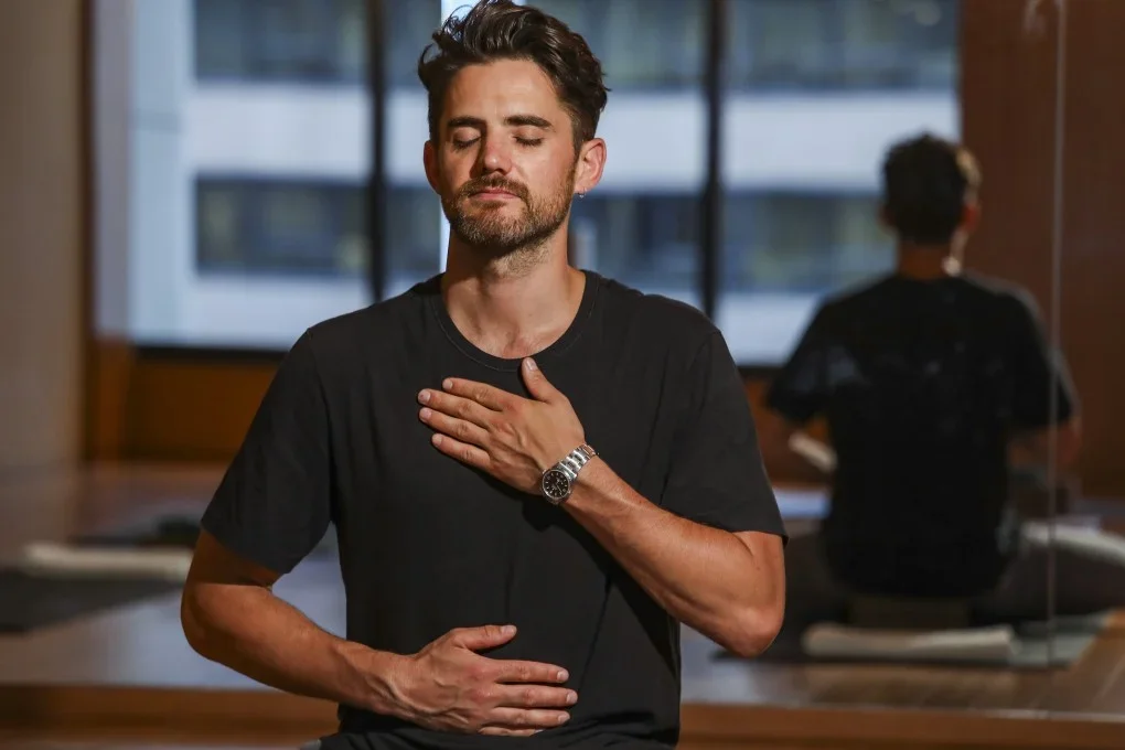 A young man with dark hair and beard practicing yoga or meditation indoors, eyes closed, one hand on his chest and the other on his stomach, in a room with large windows.