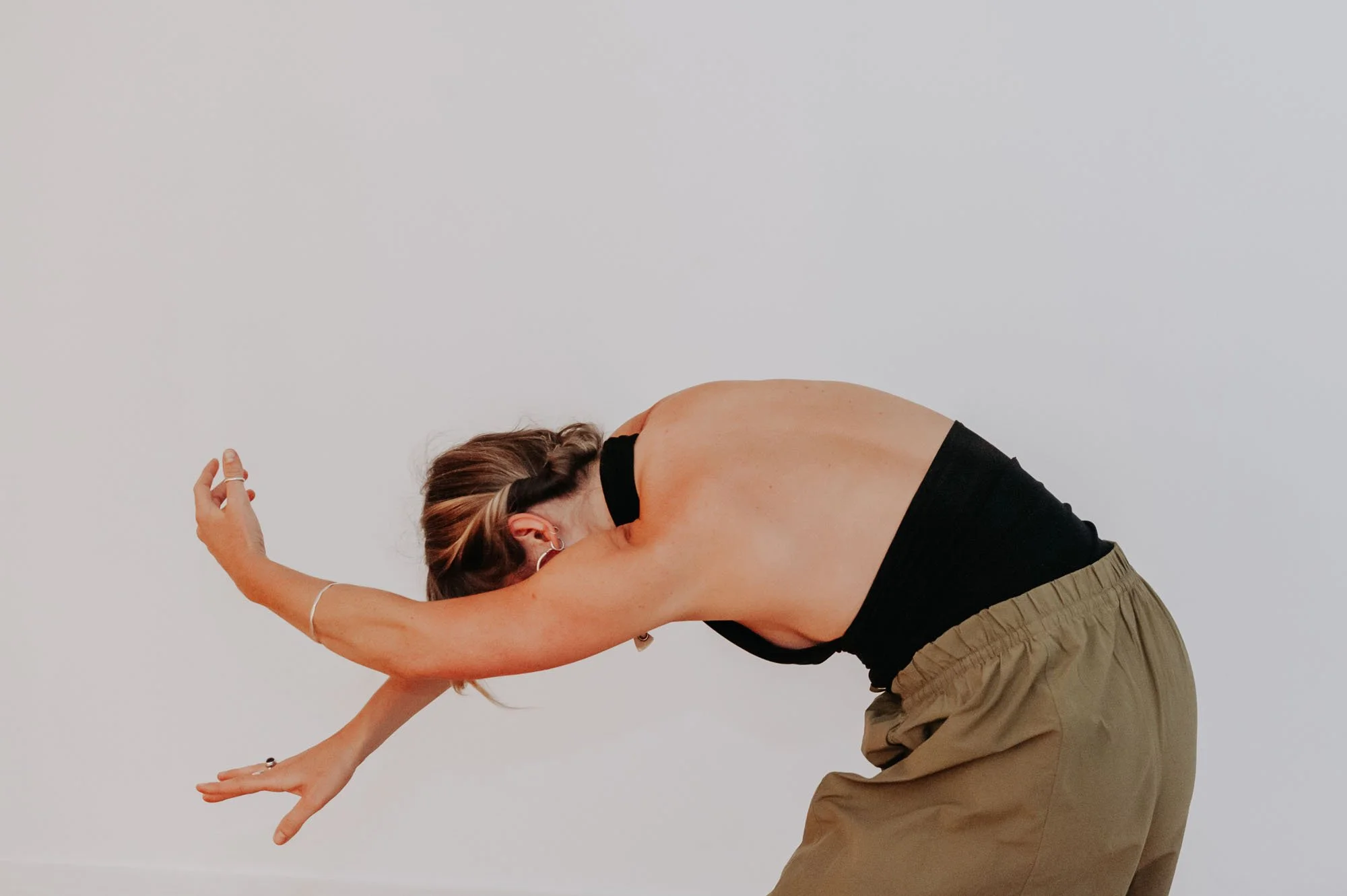 Person with short hair practicing yoga in a forward bend pose, wearing a black sleeveless top and beige pants, against a plain white background.