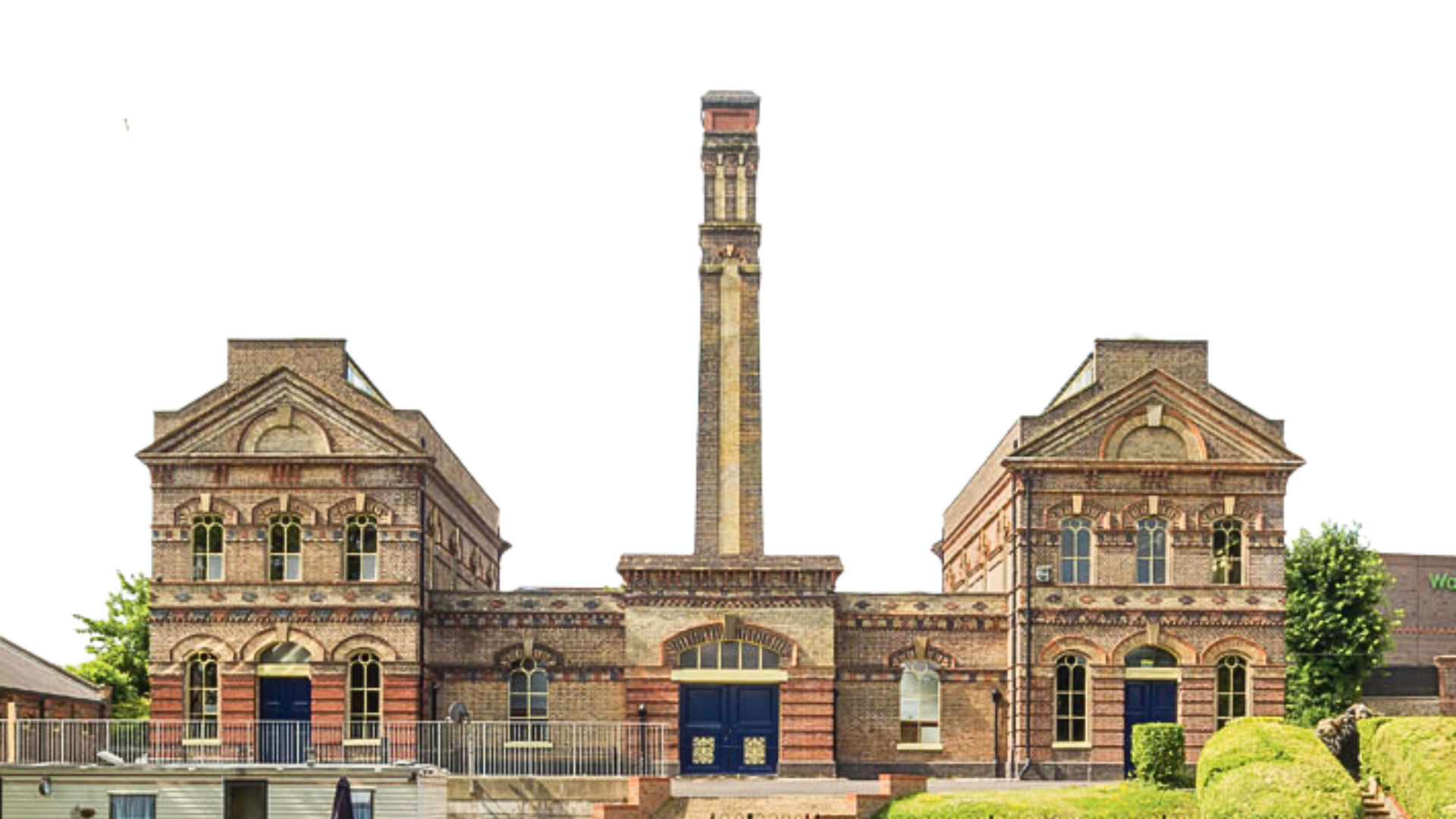 Historic brick building with a central chimney, two symmetrical wings, and arched windows, surrounded by a fence and greenery.