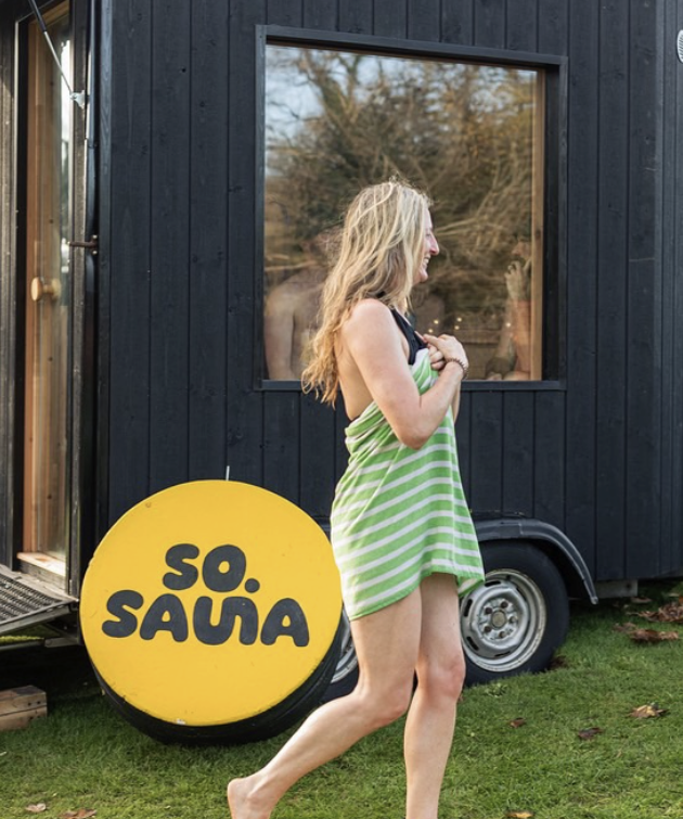 A woman in a green and white striped towel walking outdoors with a smile, near a tiny black house and a sign that says 'SO. SAUNA'.