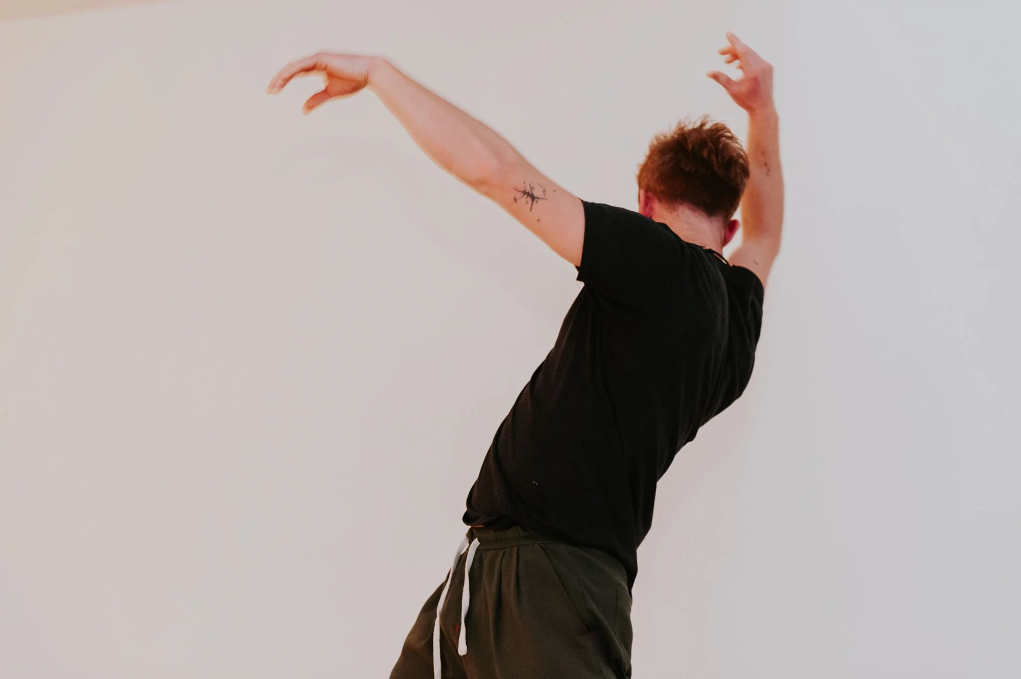 Person with short hair wearing black t-shirt and gray sweatpants stretching with arms raised against a plain white wall.