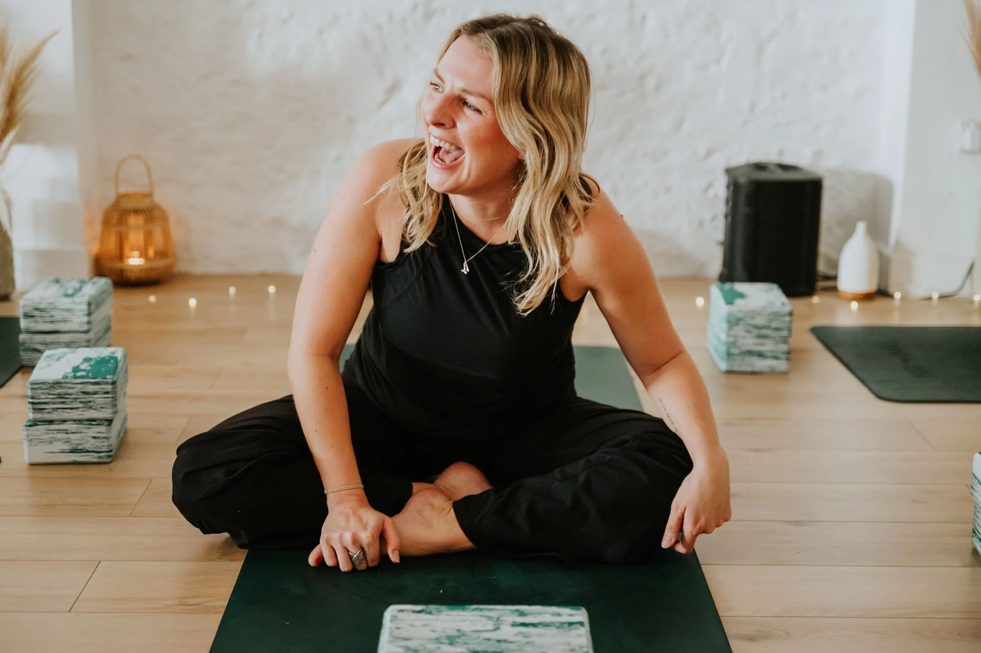 A woman with blonde hair, wearing a black sleeveless top and black pants, sitting on a yoga mat, smiling and laughing during a yoga or meditation class in a room with wooden flooring, plants, and candles.