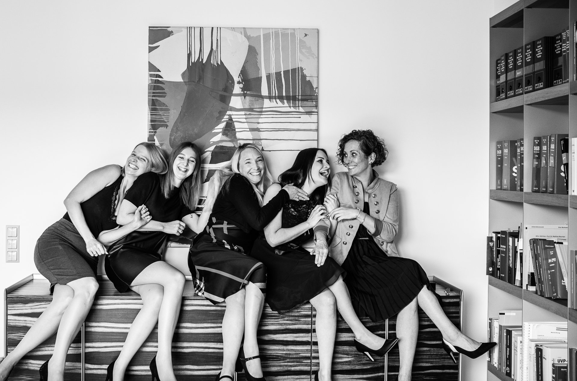 Fünf Frauen in einem Büro lachen und sitzen auf einer Anrichte vor einem abstrakten Gemälde. Im Hintergrund sind Regale mit Büchern.