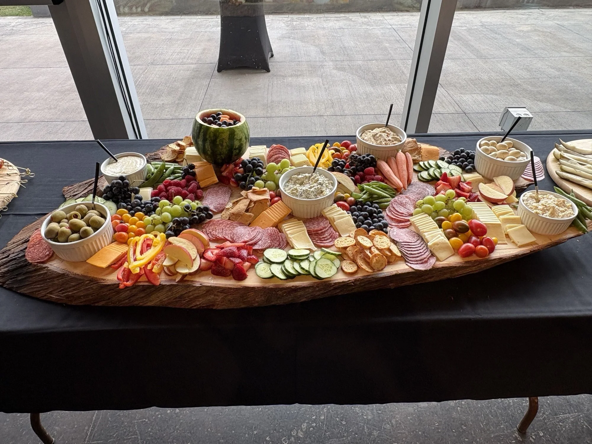 A cheese and fruit platter on a wooden board with bowls of dip or yogurt, sliced meats, grapes, watermelon, strawberries, cucumber, cherry tomatoes, and sliced cheese, set on a black table near a window.
