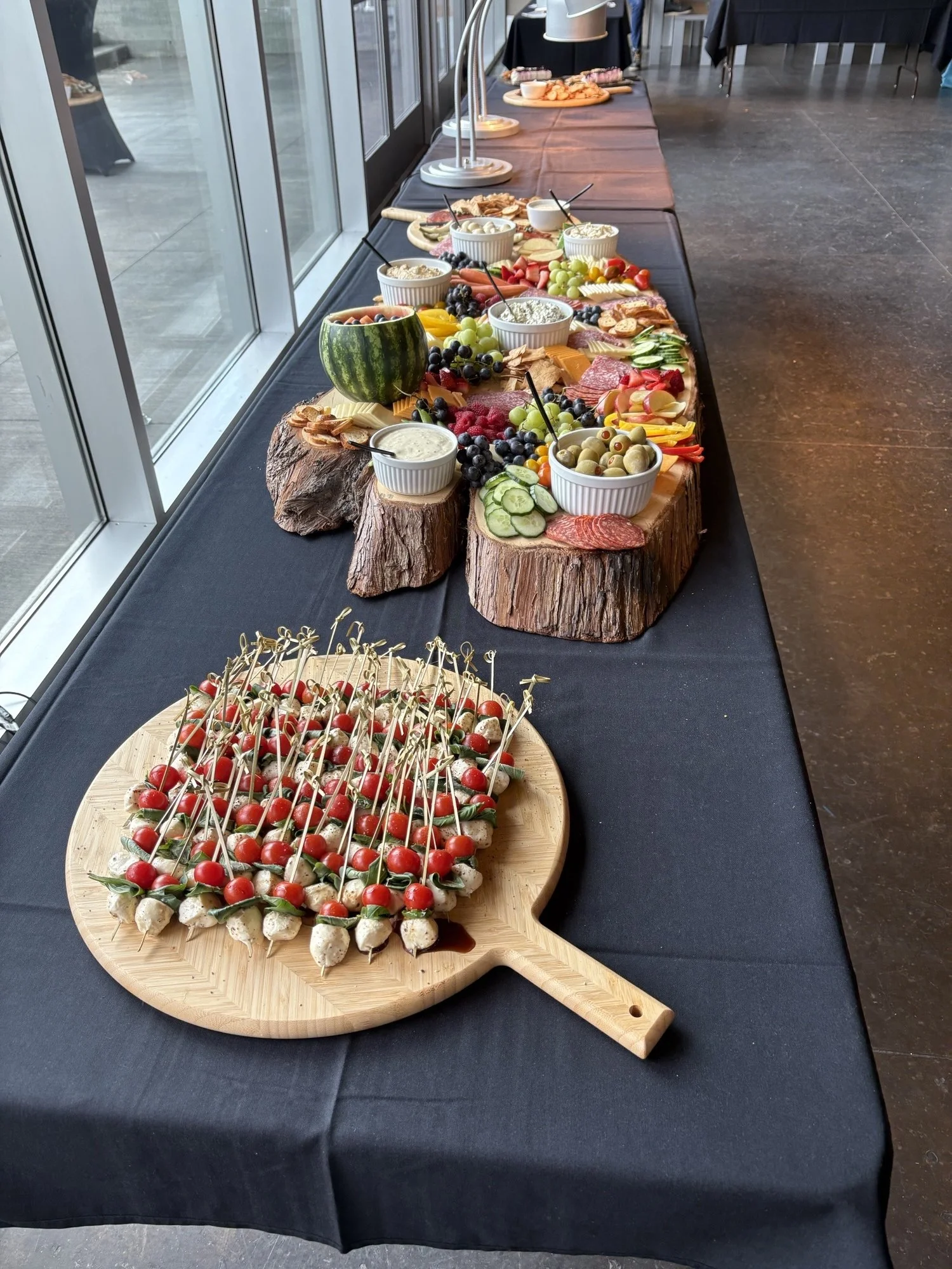 A long table with black tablecloth displaying a spread of food, including bowls of dips and a carved watermelon, with an assortment of fruits, vegetables, meats, and cheese, all arranged on wooden and stone slabs in a banquet hall.