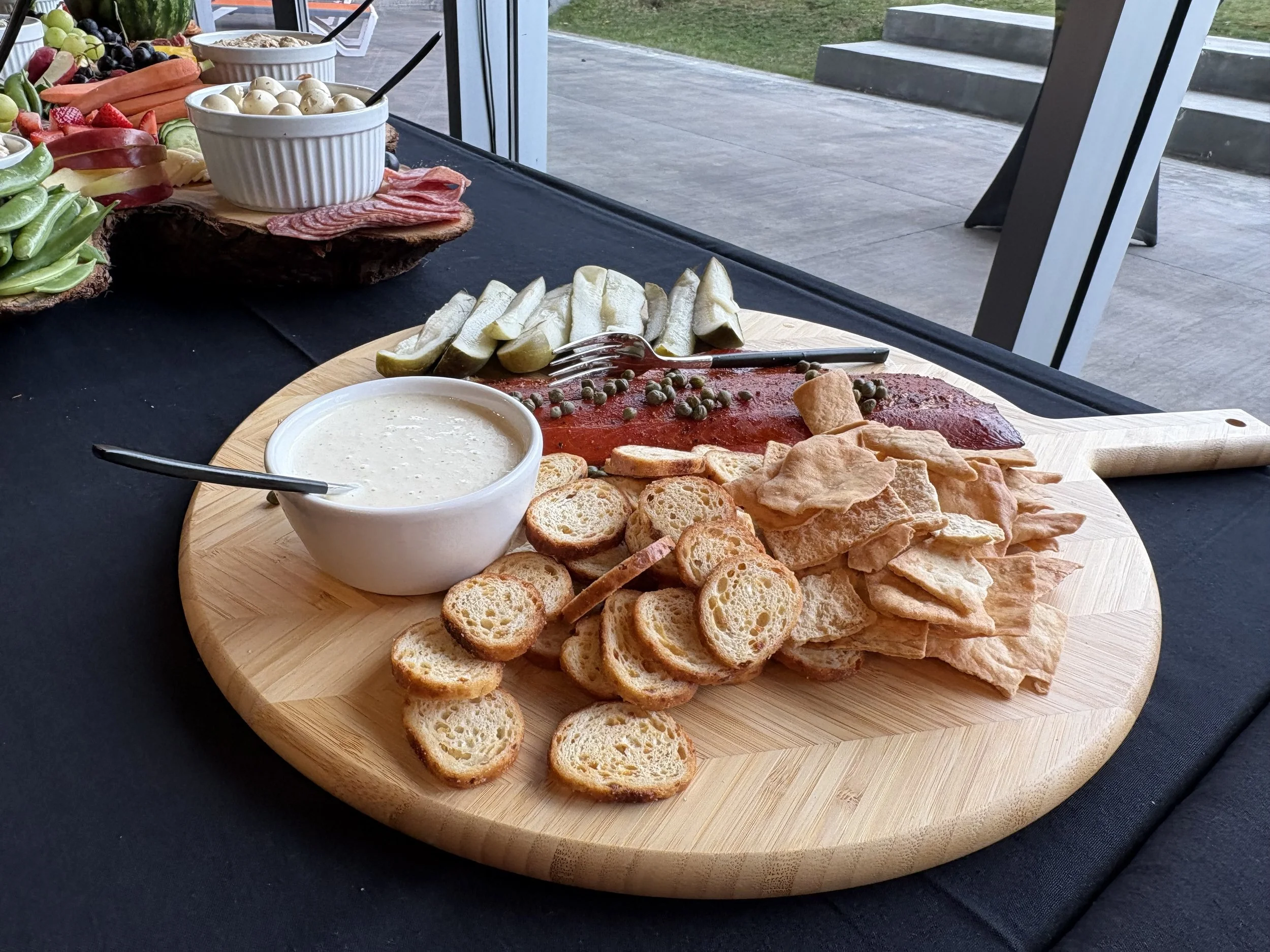 A round wooden serving board with sliced baguette bread, pickles, capers, and a small bowl of creamy dipping sauce on a black table.