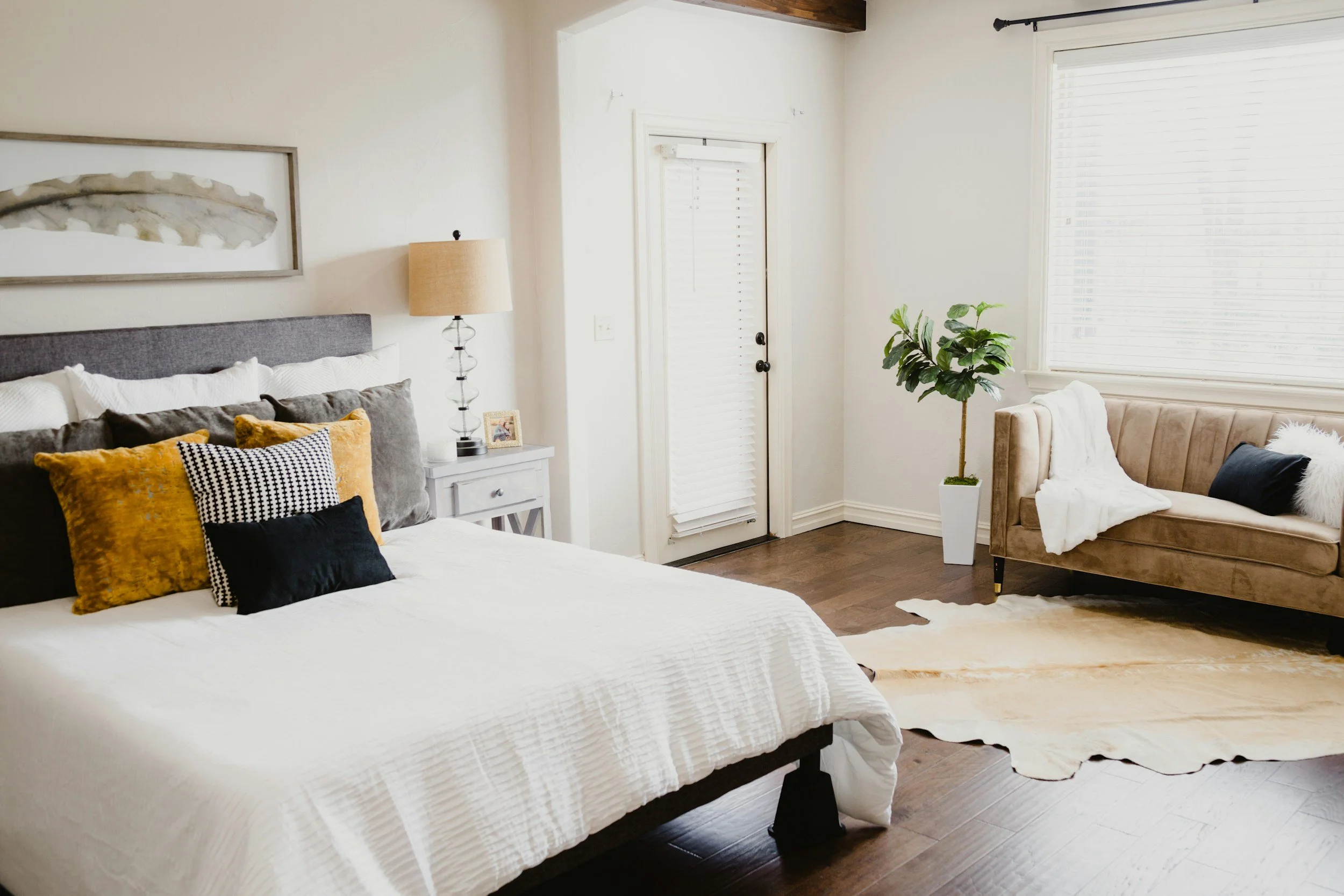 A bedroom with a bed on the left, a white nightstand with a lamp, and a small picture frame. To the right, there's a beige sofa with pillows and a throw blanket, next to a large window with blinds and a potted plant. The room has wooden floors and a cowhide rug.