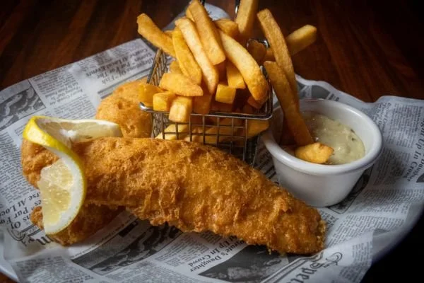 Fried fish fillet with lemon wedge, French fries in a basket, and tartar sauce on newspaper-style paper.