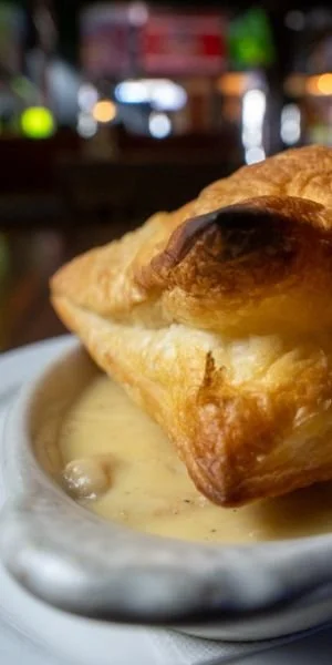 Close-up of a golden-brown pastry on a white dish with creamy dipping sauce, blurred restaurant background.