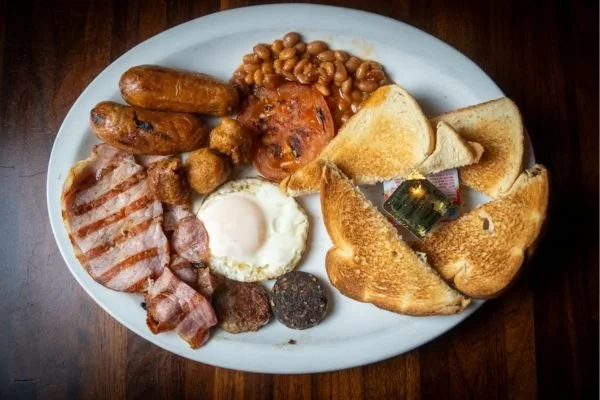 Full English breakfast with fried eggs, bacon, sausage, baked beans, toast, and hash browns on a white plate.