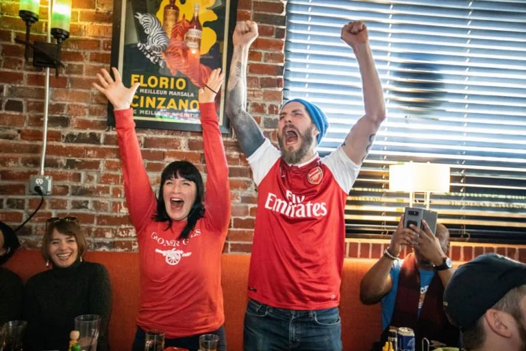 Group of people cheering in a restaurant, one wearing an Arsenal jersey and the other in a red shirt, with arms raised in celebration.