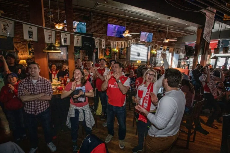 A group of people in a pub cheering and watching a sports game on televisions. Some wear team jerseys and scarves, and the space is decorated with sports memorabilia.