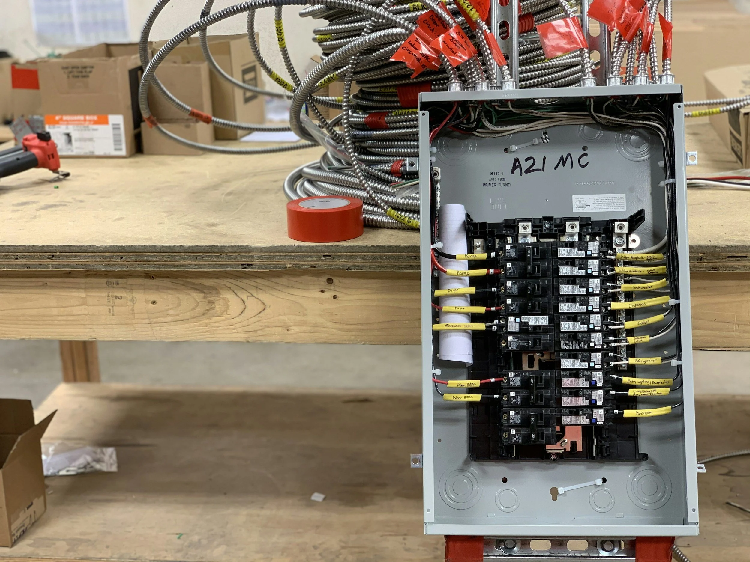 Open electrical panel with multiple labeled wires and circuit breakers, with a workbench and tools in the background.