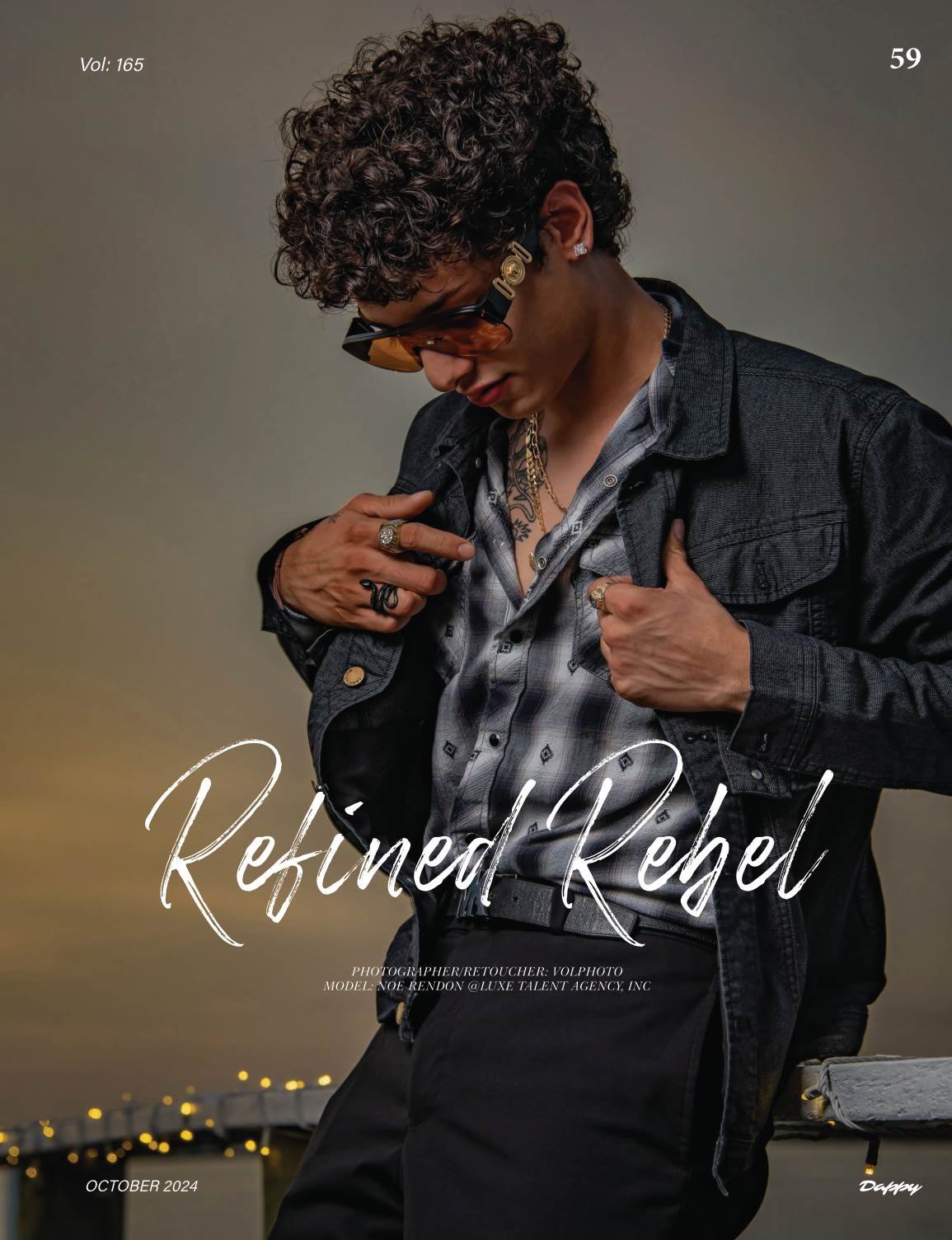 A young man with curly hair, sunglasses, and jewelry, wearing a patterned shirt and black denim jacket, posing against a sunset background with city lights.