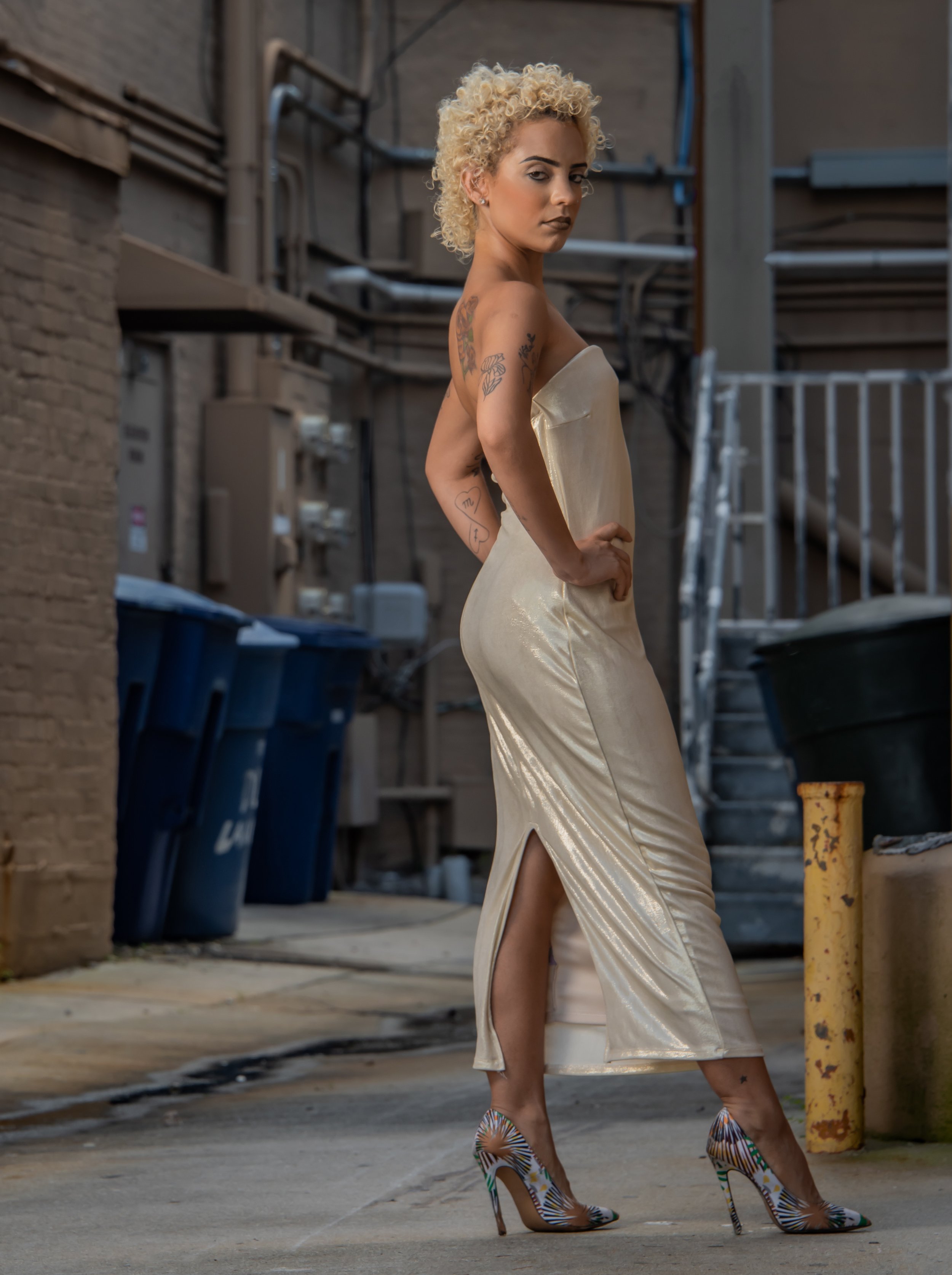 Portrait of a blonde curly haired female model posting in an alley wearing a light gold shimmery dress and heels posing with her hands on her hips.