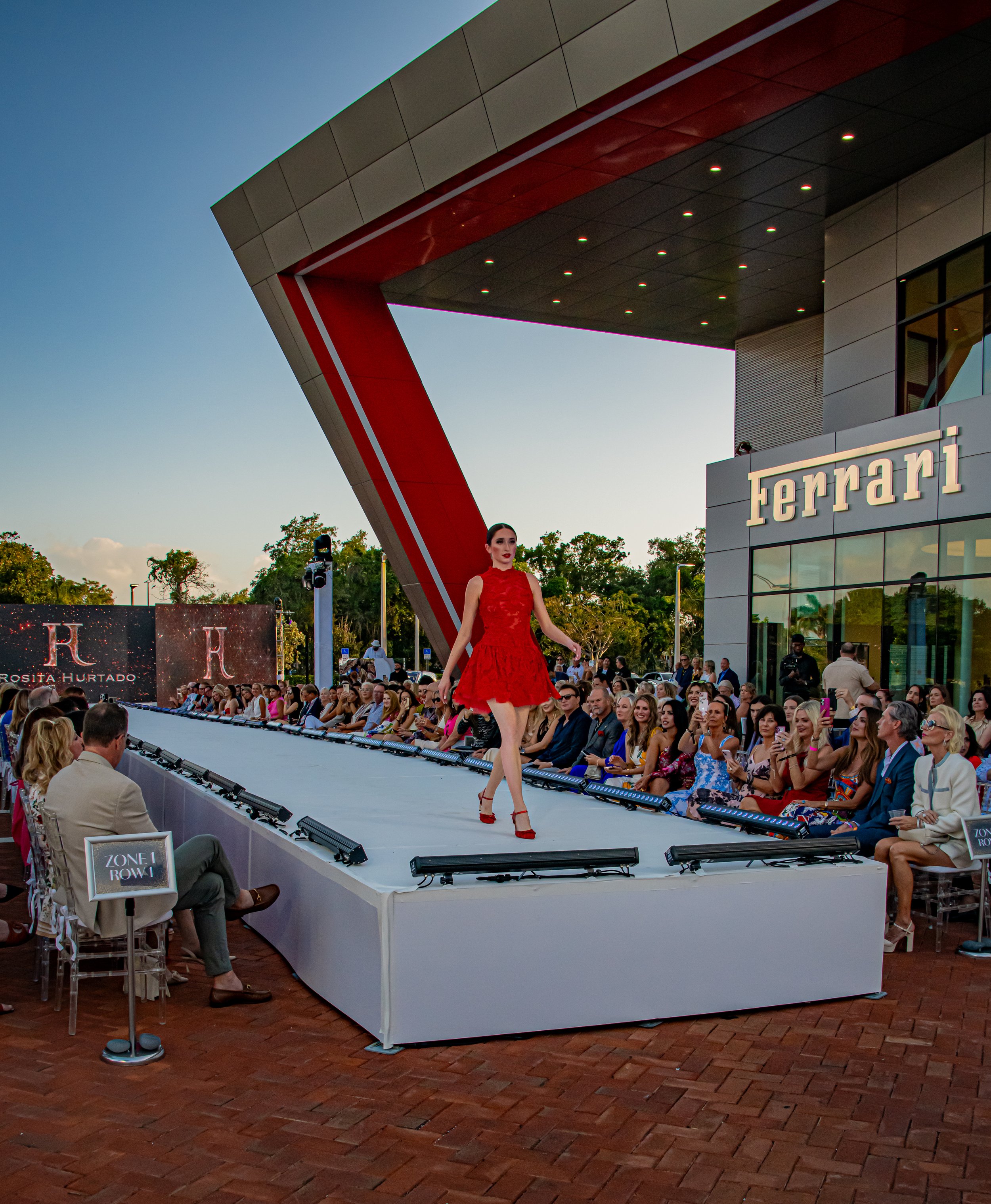 A fashion runway show at a Ferrari dealership with a model in a red dress walking on the runway, and an audience watching.